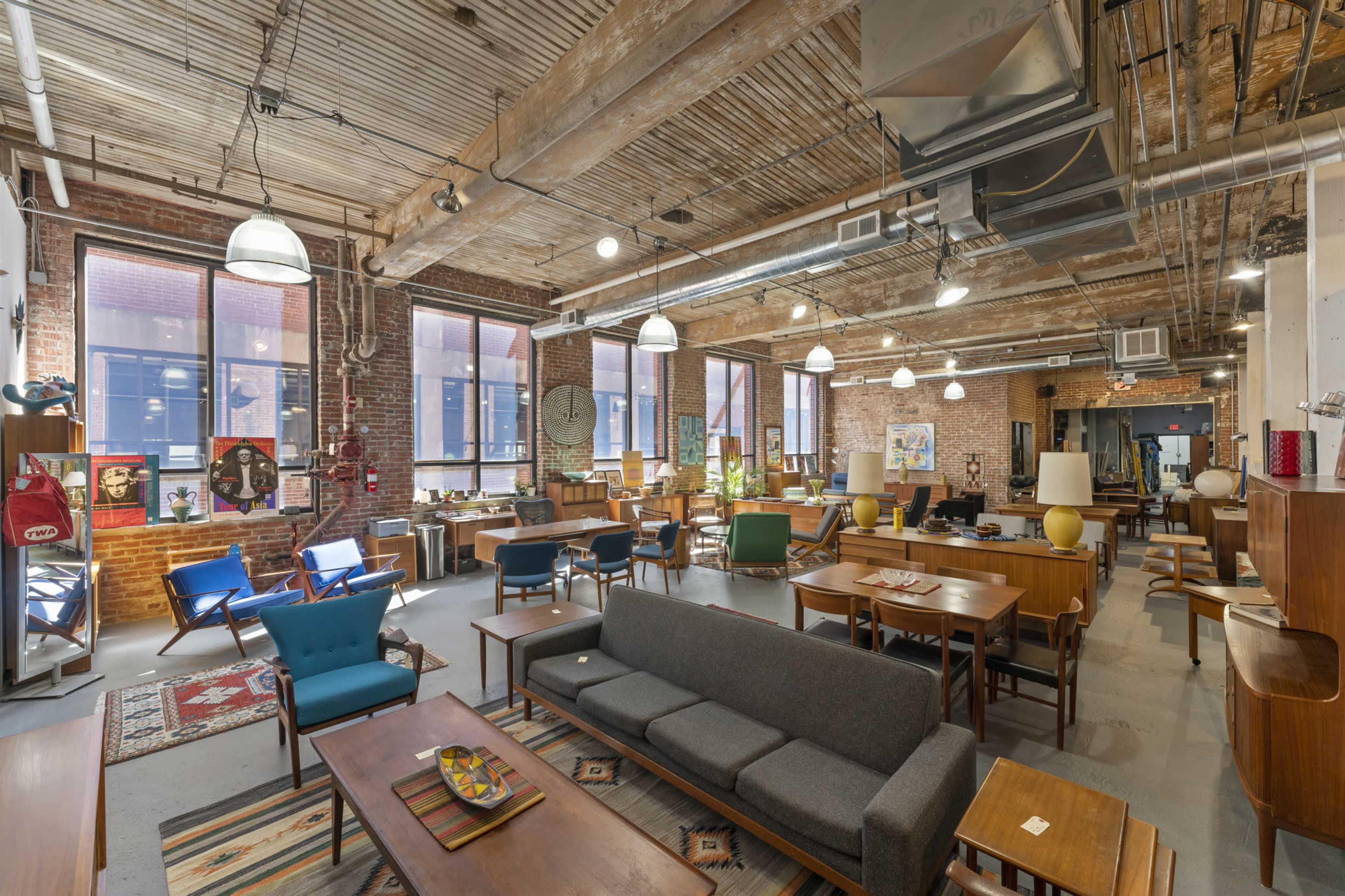 A spacious, well-lit room filled with various vintage furniture pieces, including sofas, chairs, and tables, against a backdrop of exposed brick walls and large windows.