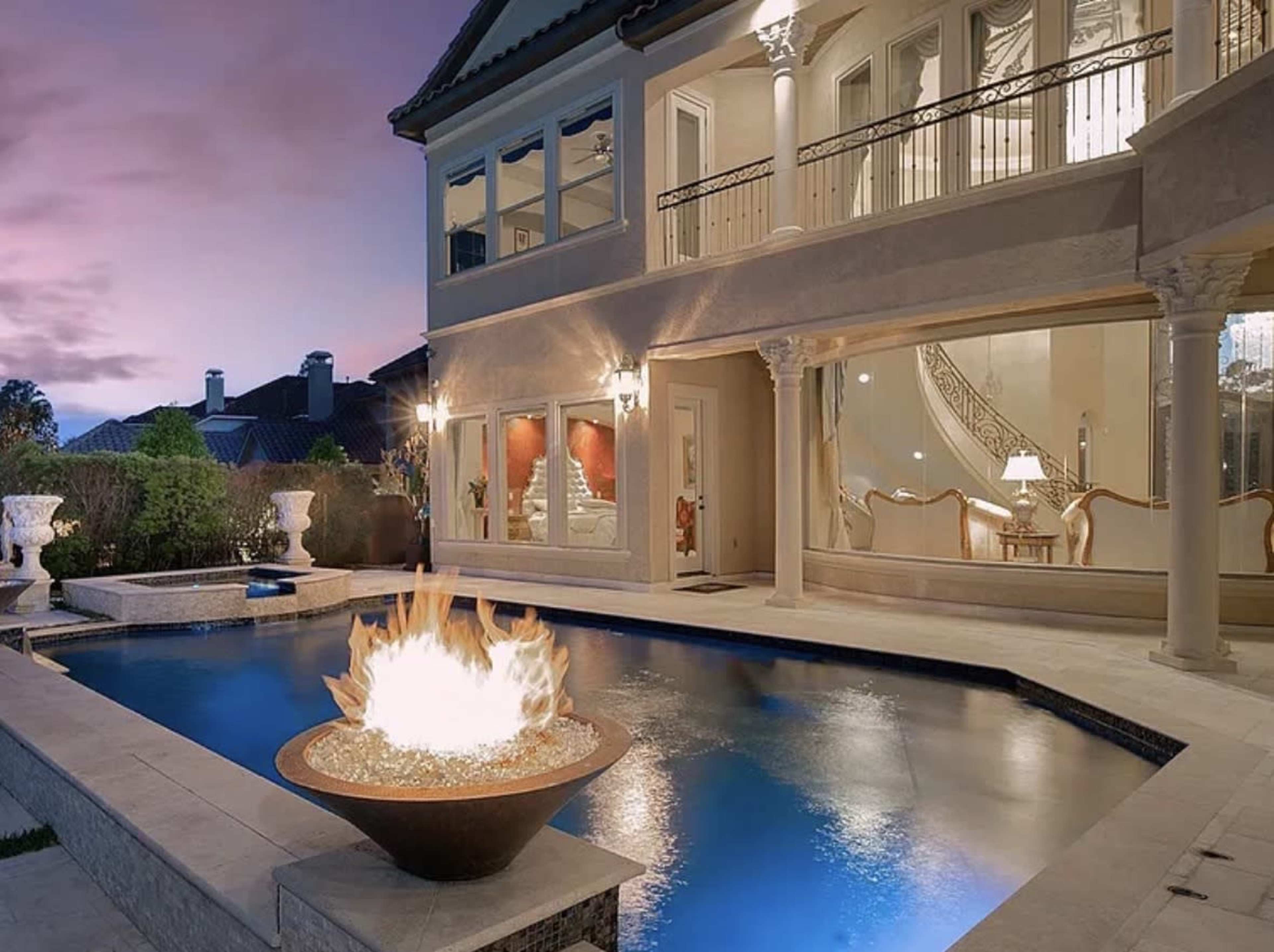 A luxurious backyard features a swimming pool, an outdoor fire bowl, and an elegant two-story house with large windows.