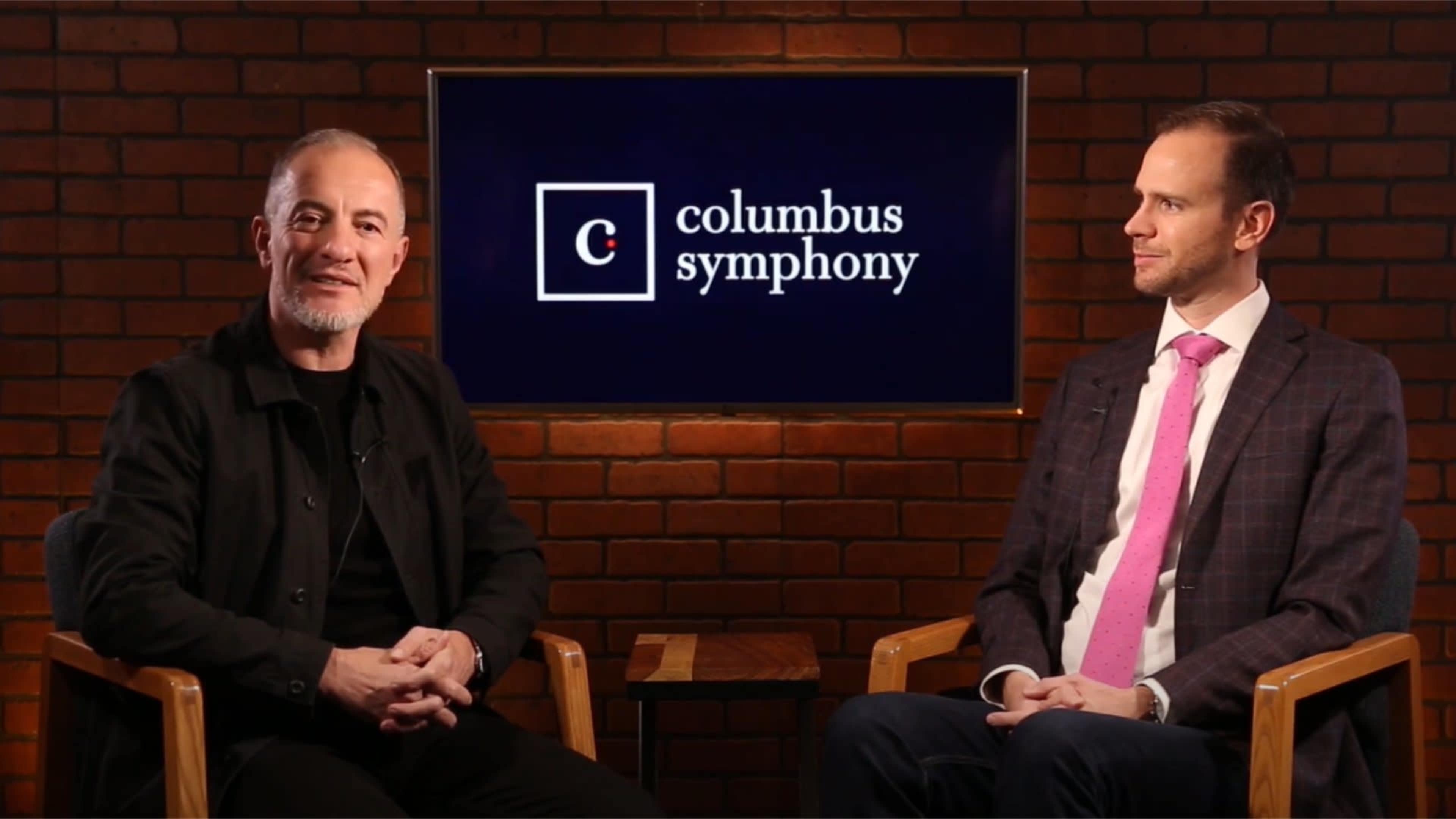 Two men sit in chairs facing each other in a studio setting, with a brick wall in the background and a sign displaying "Columbus Symphony" prominently behind them.