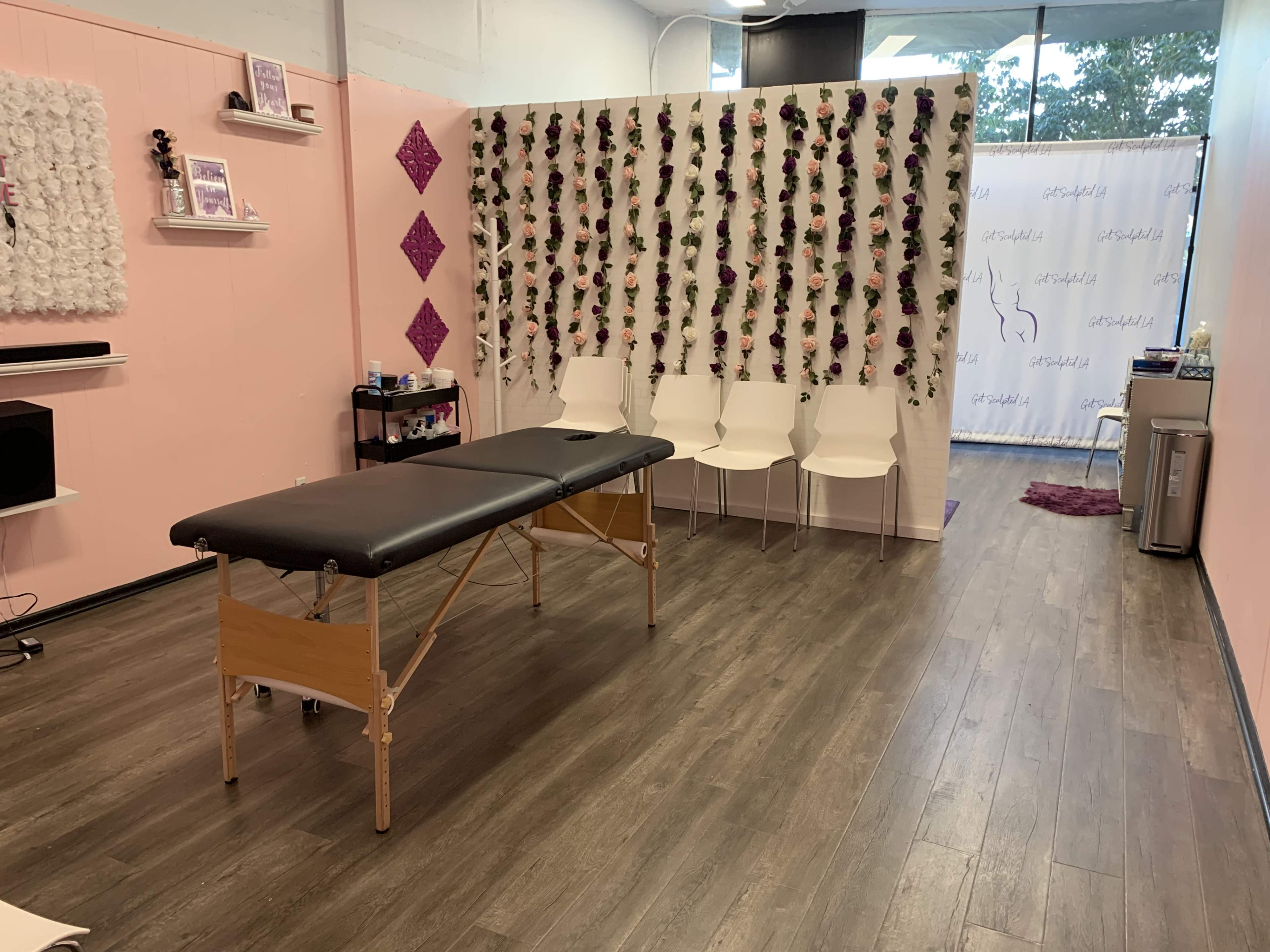 A treatment room with a massage table in the center, surrounded by pink walls and decorative elements, including a wall adorned with artificial flowers.