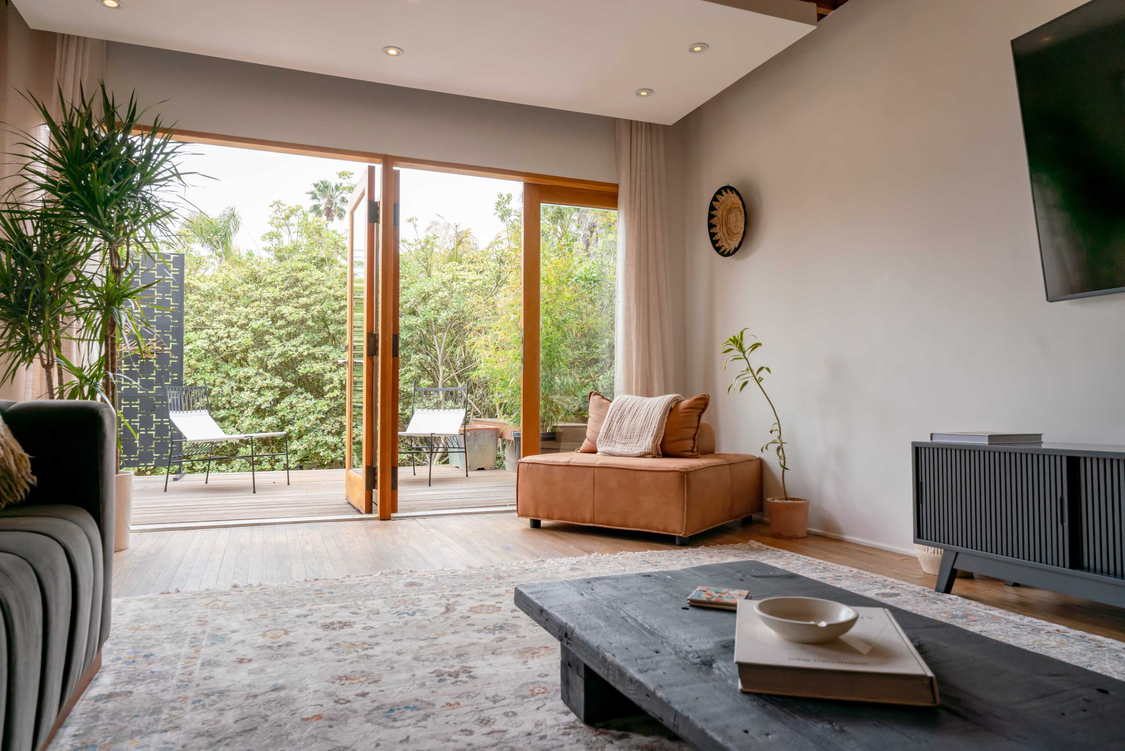 The image shows a modern living room with a brown leather couch, a wooden coffee table, and large sliding doors that open onto a patio surrounded by greenery.