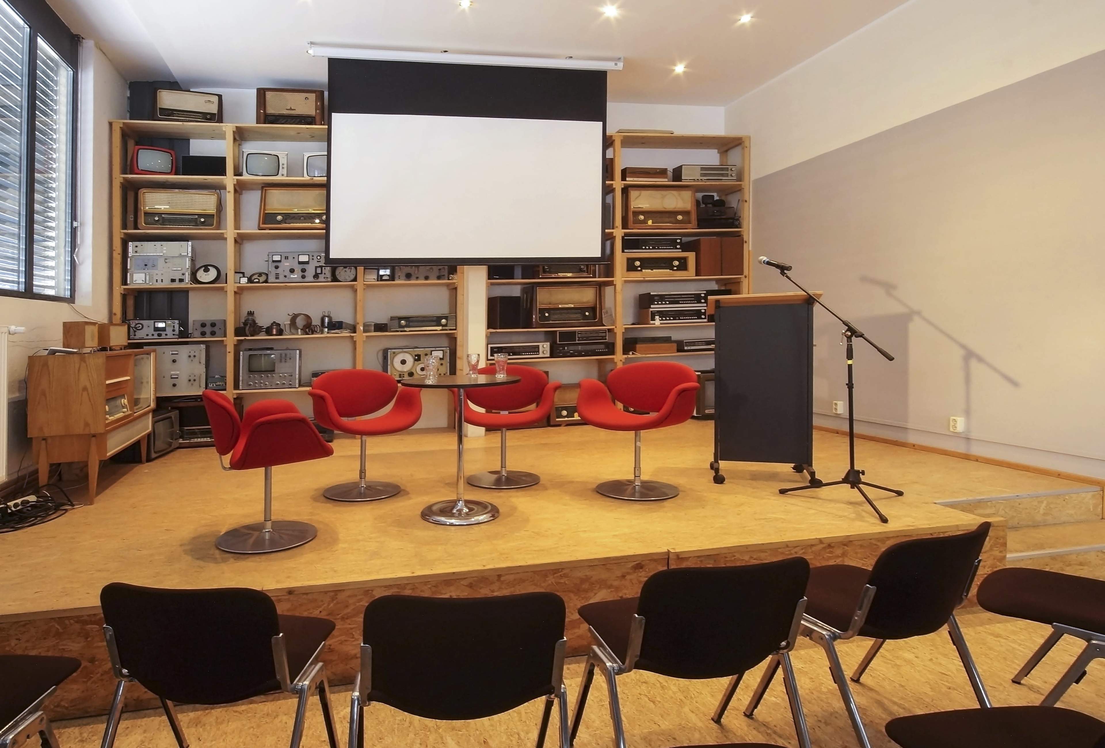 The image shows a small auditorium setup with four red chairs around a circular table, a podium with a microphone, and a large projector screen against a backdrop of vintage audio equipment.