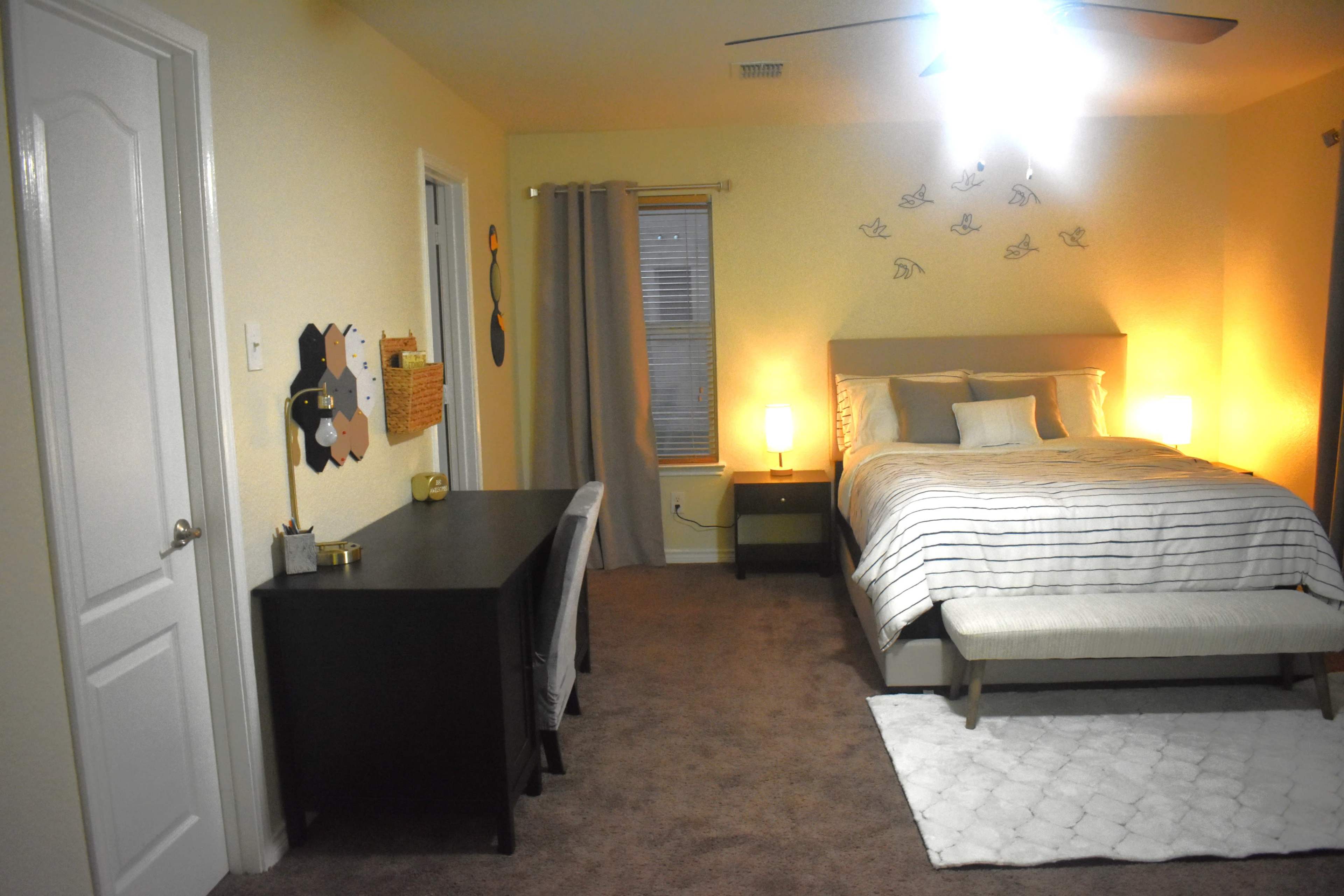 The image shows a bedroom with a bed, a desk, and warm lighting, featuring beige walls and carpeted flooring.