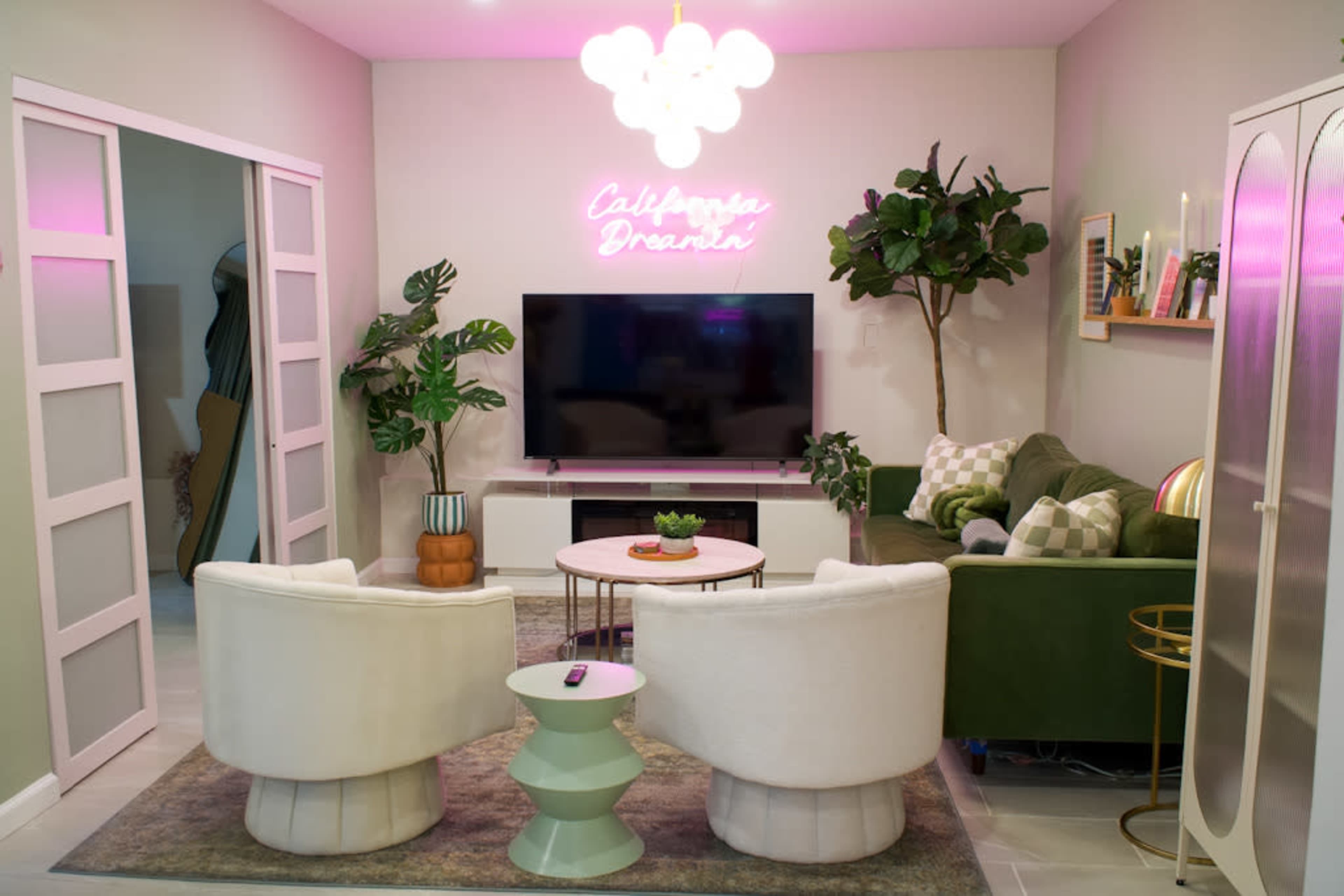 A modern living room features a green sofa, two white armchairs, a large television, and decorative plants, with a neon sign that reads "California Dreamin'."