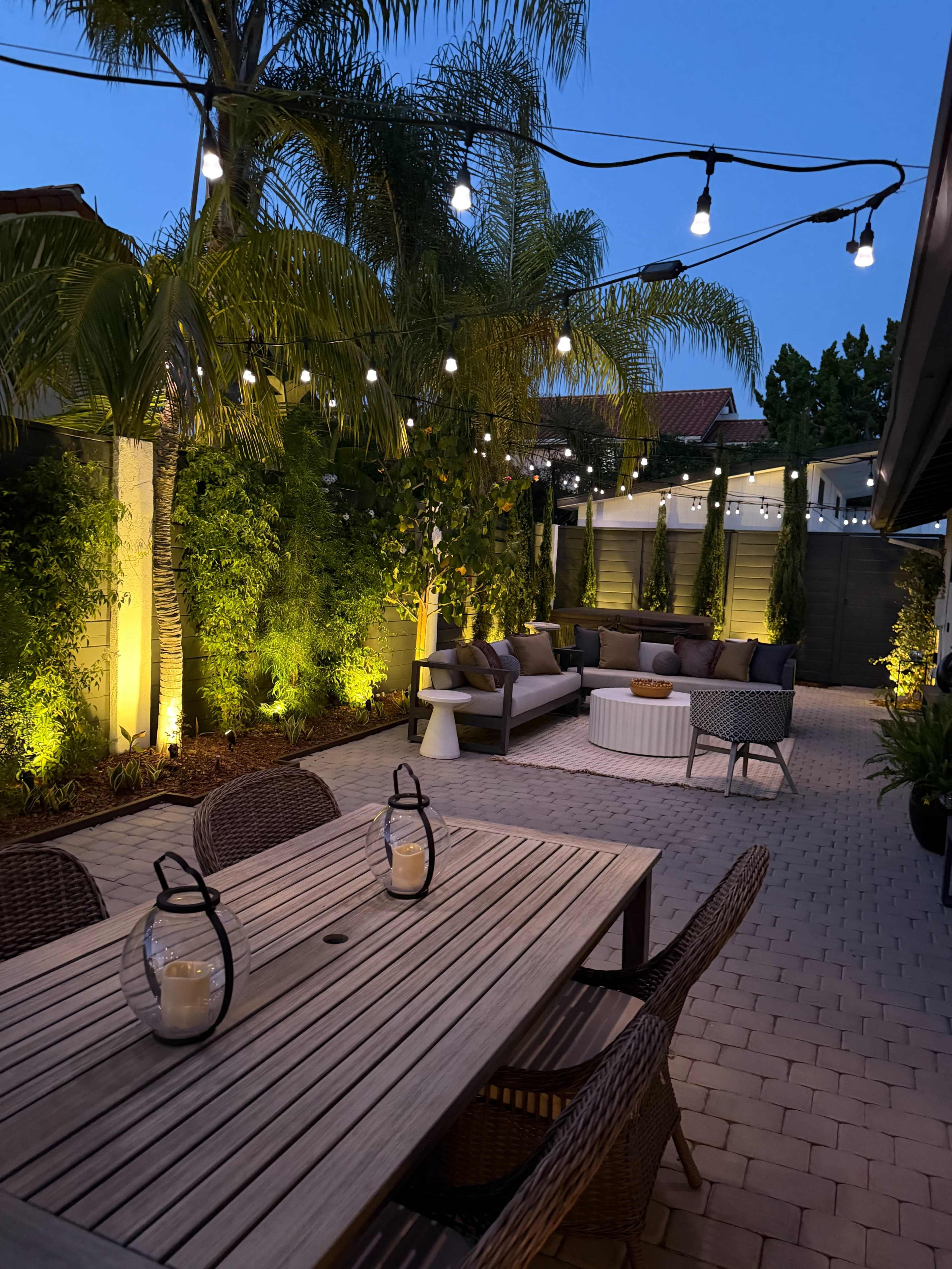 The image shows a landscaped outdoor patio area with string lights, potted palm trees, a seating arrangement, and a wooden dining table with lanterns.
