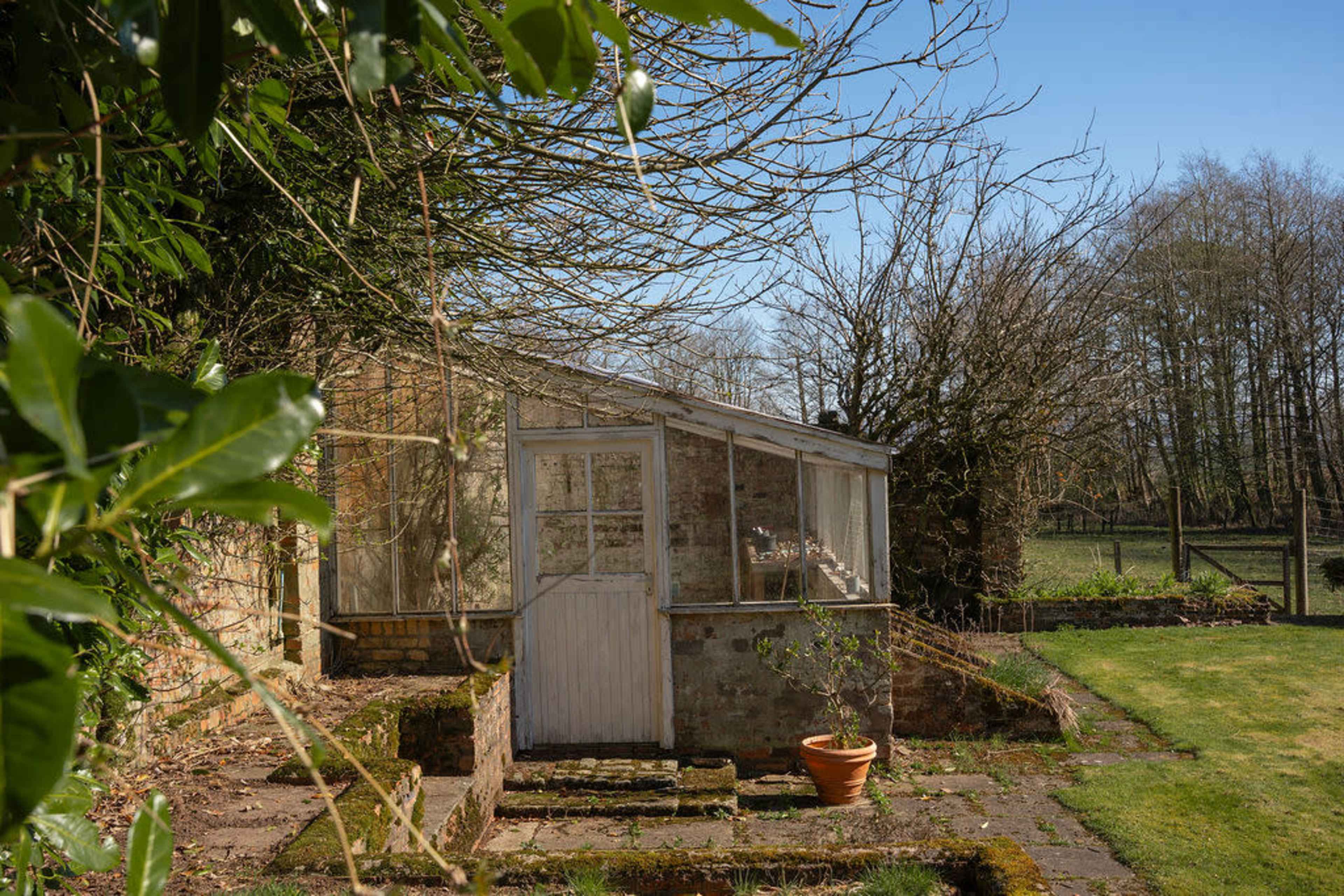 A small greenhouse with a white door and large windows is situated beside a brick wall, surrounded by grass and trees.