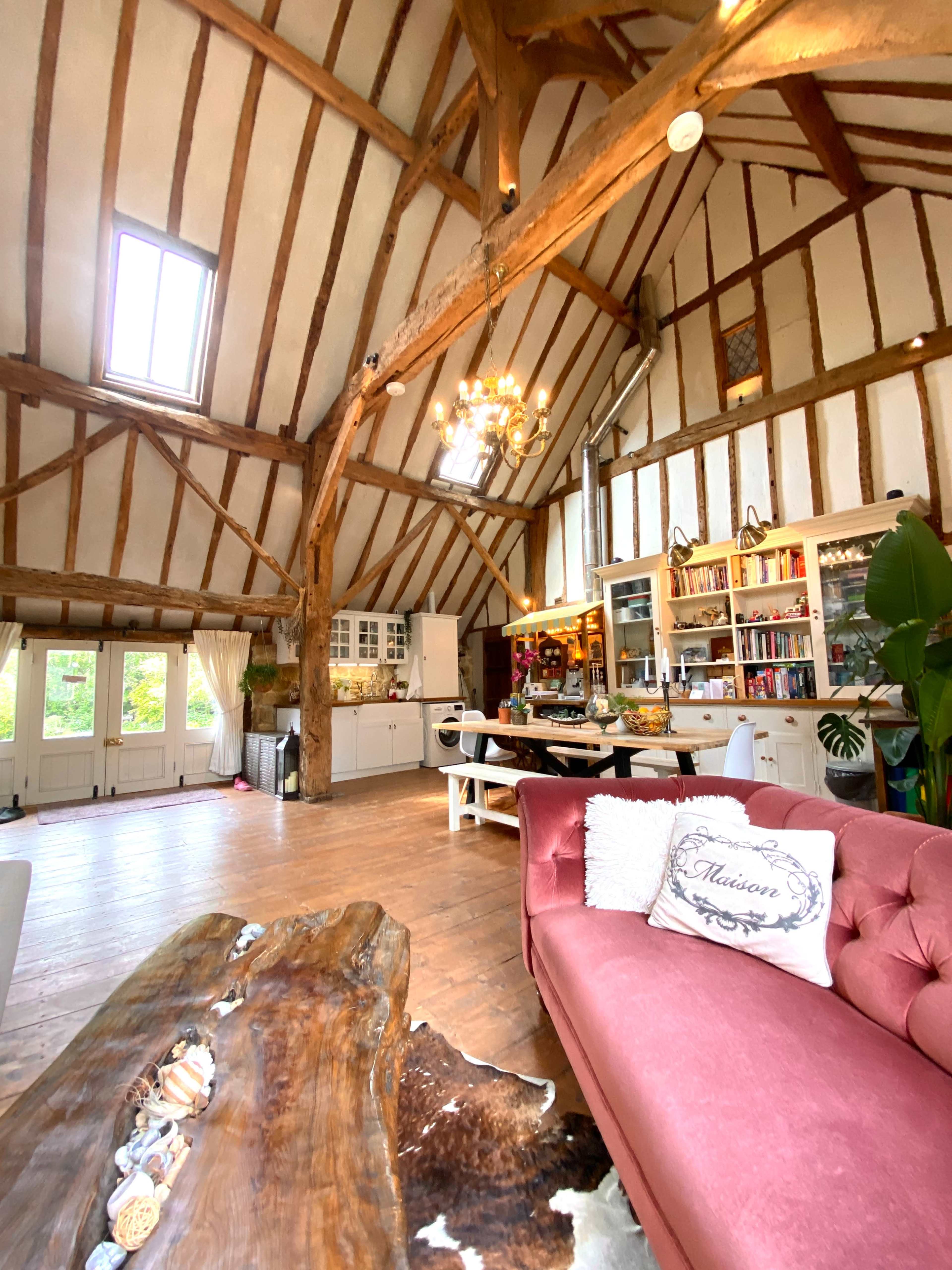 A spacious, rustic living area with wooden beams, a large dining table, a pink sofa, and a kitchen area in a bright, well-lit room.