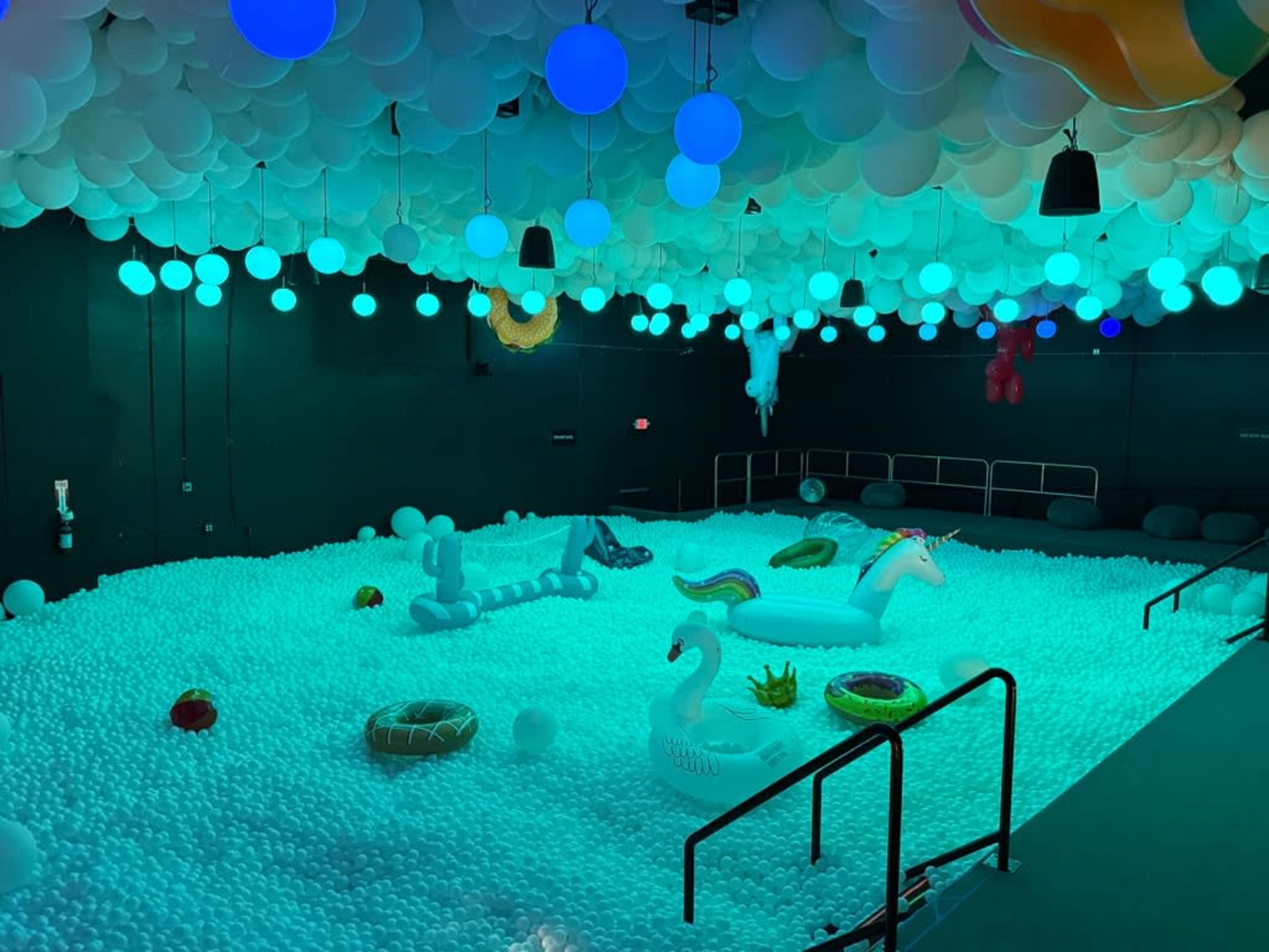 The image shows a large room filled with colorful inflatable toys floating on a sea of white plastic balls, illuminated by blue and green lighting and decorated with multicolored hanging spheres.