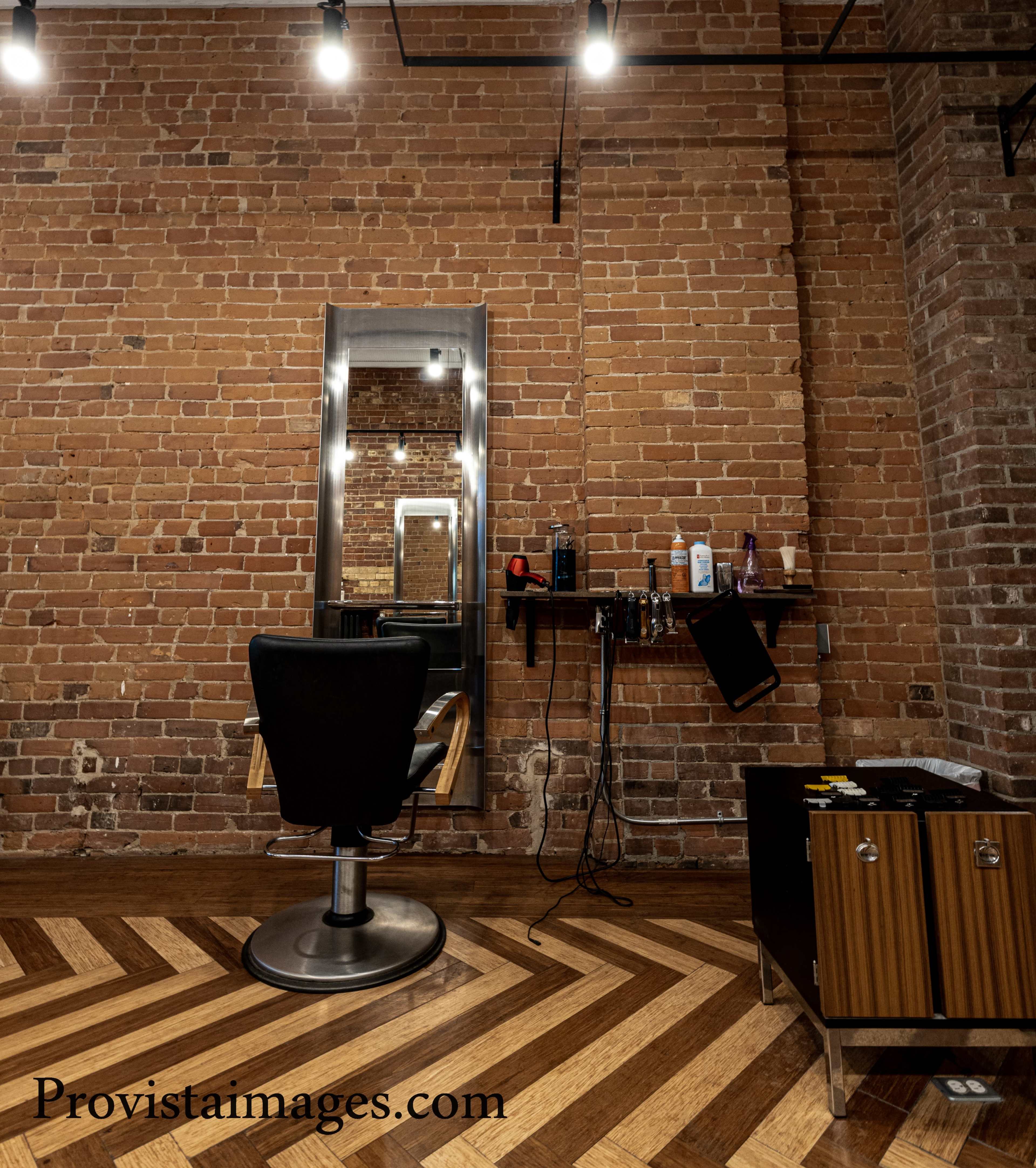 A salon interior with a barber chair facing a mirror, set against a brick wall, with wooden herringbone flooring and a shelf containing hair products.