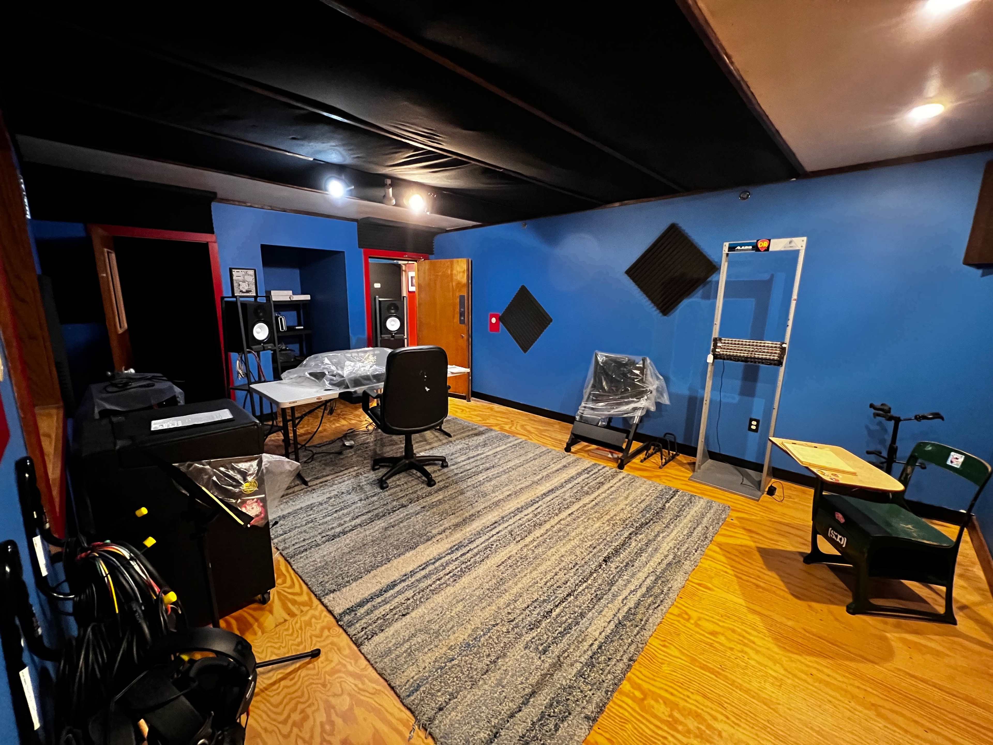 The image shows a recording studio with blue walls, a large rug on the wooden floor, and various equipment covered in plastic.