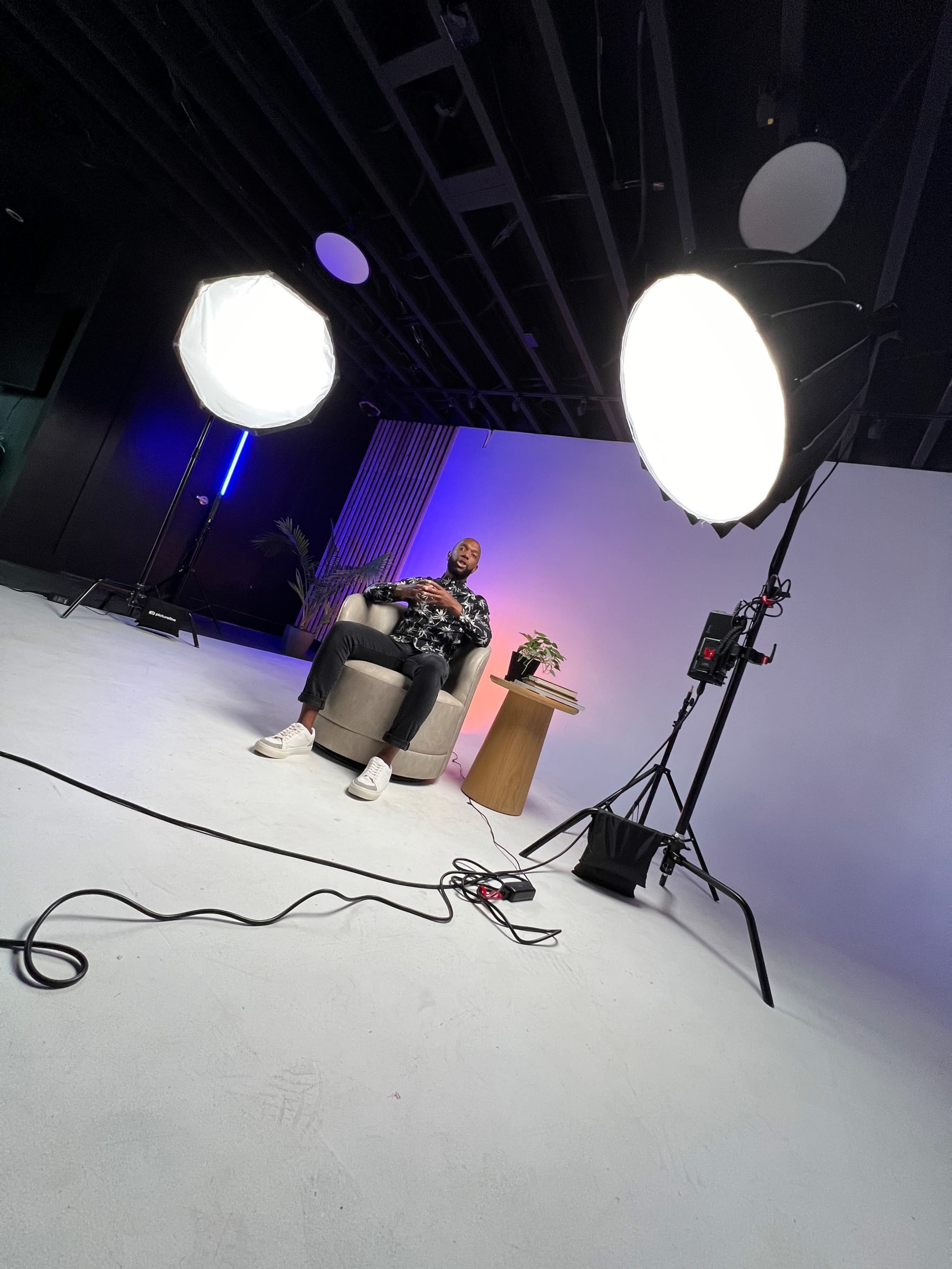 A person sits in a chair in a studio setting, flanked by two large lights and a small table with a potted plant.