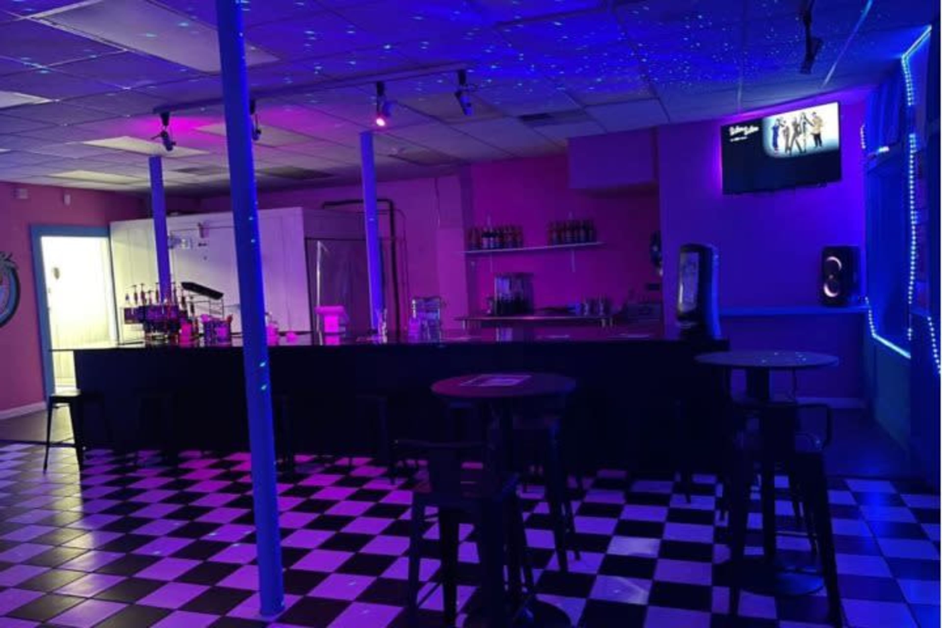 A dimly lit bar area with a checkered floor, a black countertop, and colorful lighting effects.
