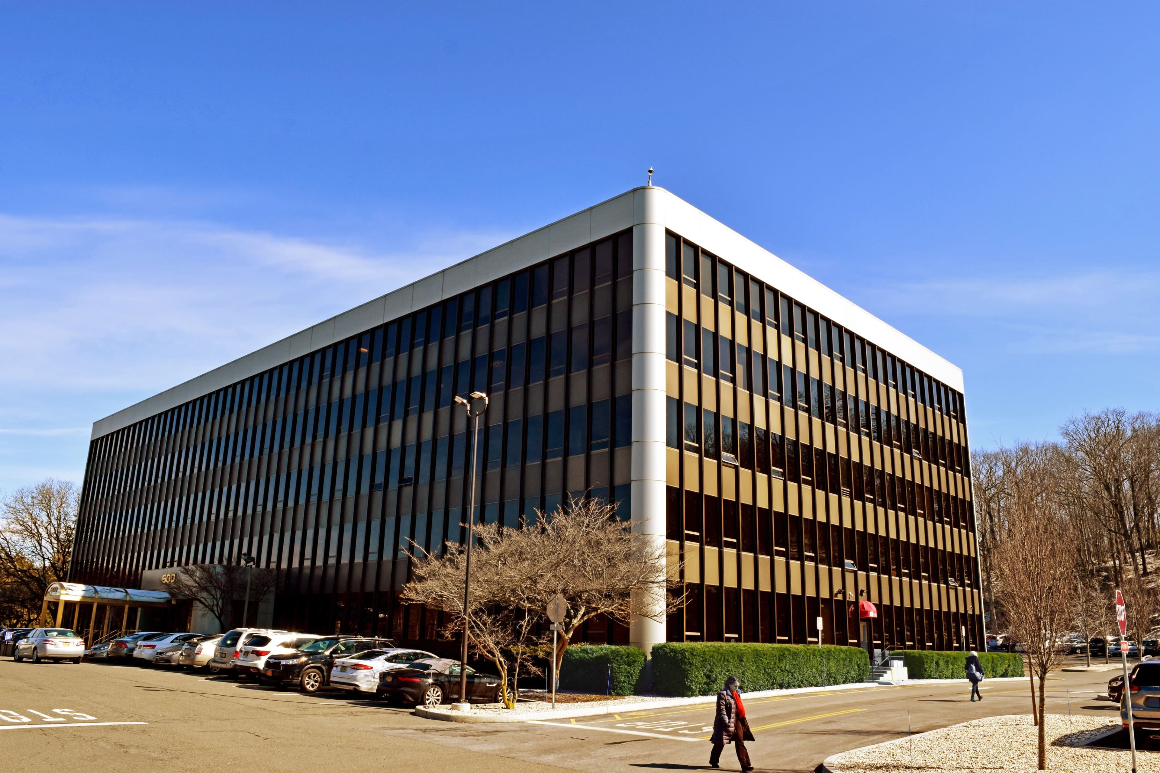 A modern, five-story office building with a glass facade stands beside a parking lot and pedestrian walkway under a clear blue sky.