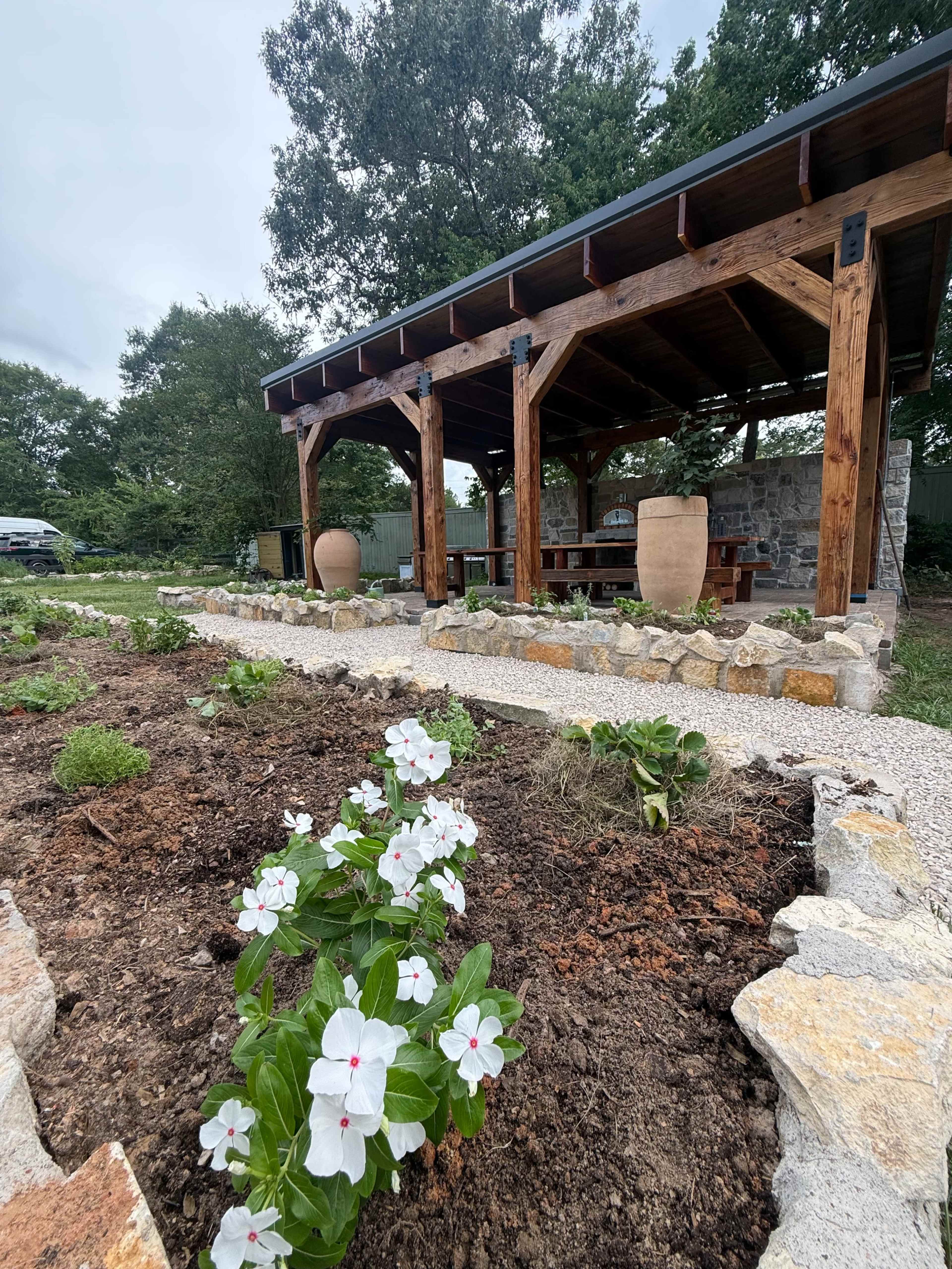 A garden area featuring a wooden pavilion with a stone foundation, surrounded by freshly tilled soil and blooming white flowers.