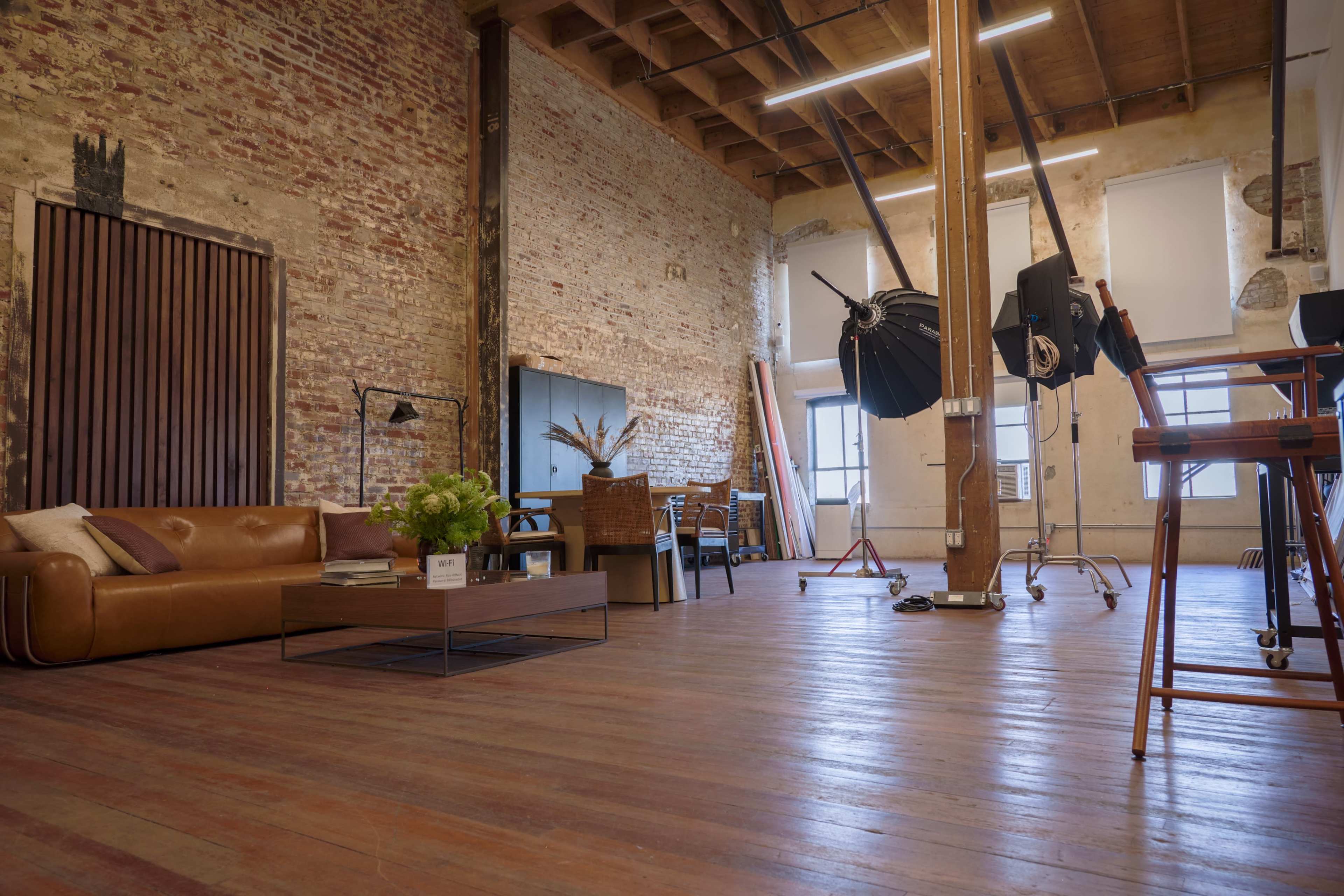 The image shows a spacious, industrial-style room with exposed brick walls, wooden beams, and a mix of furniture including a leather sofa, a coffee table, and photography equipment.