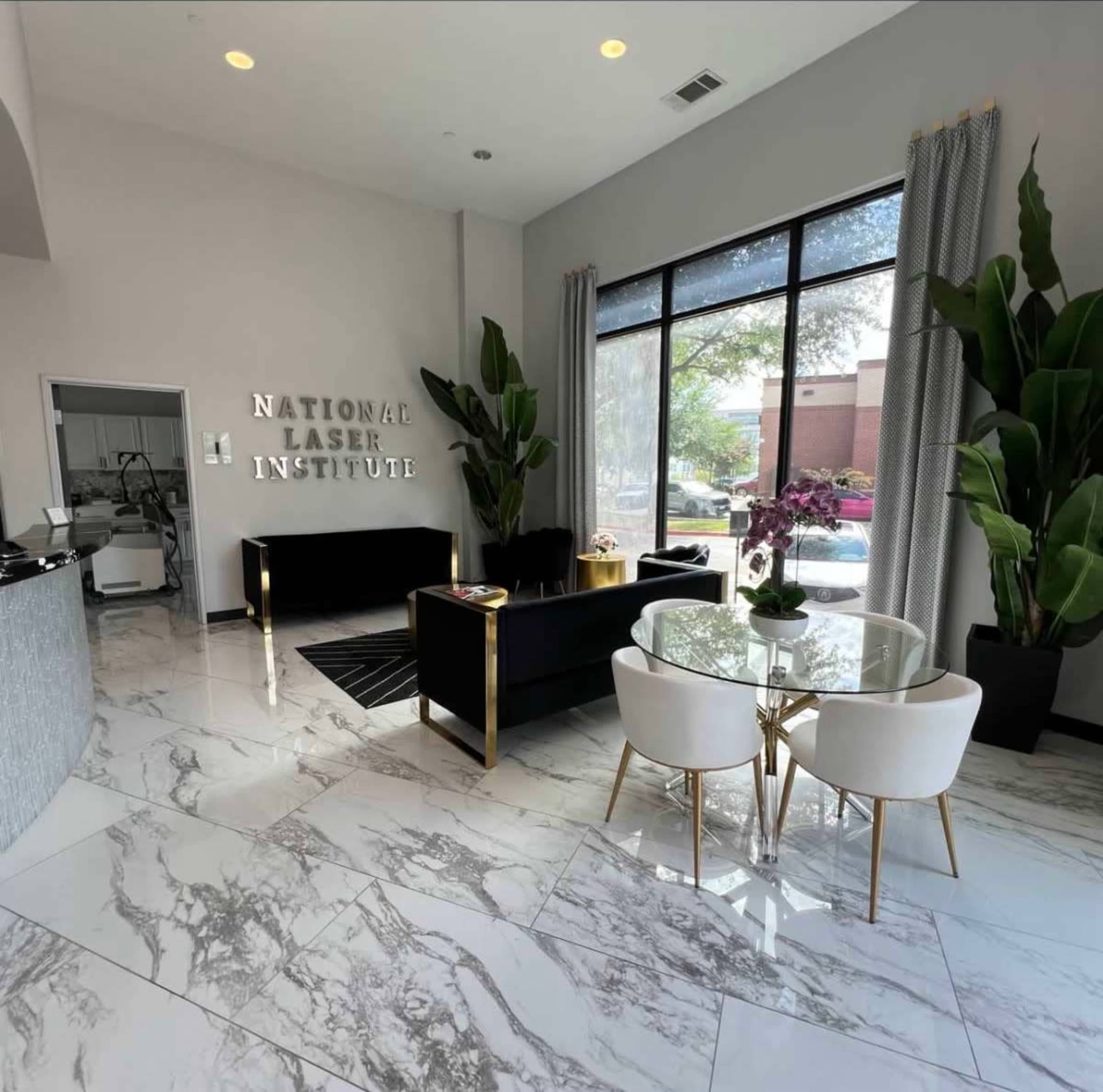 The image shows a modern reception area of the National Laser Institute featuring marble flooring, two black sofas, a round dining table with white chairs, and large windows allowing natural light.
