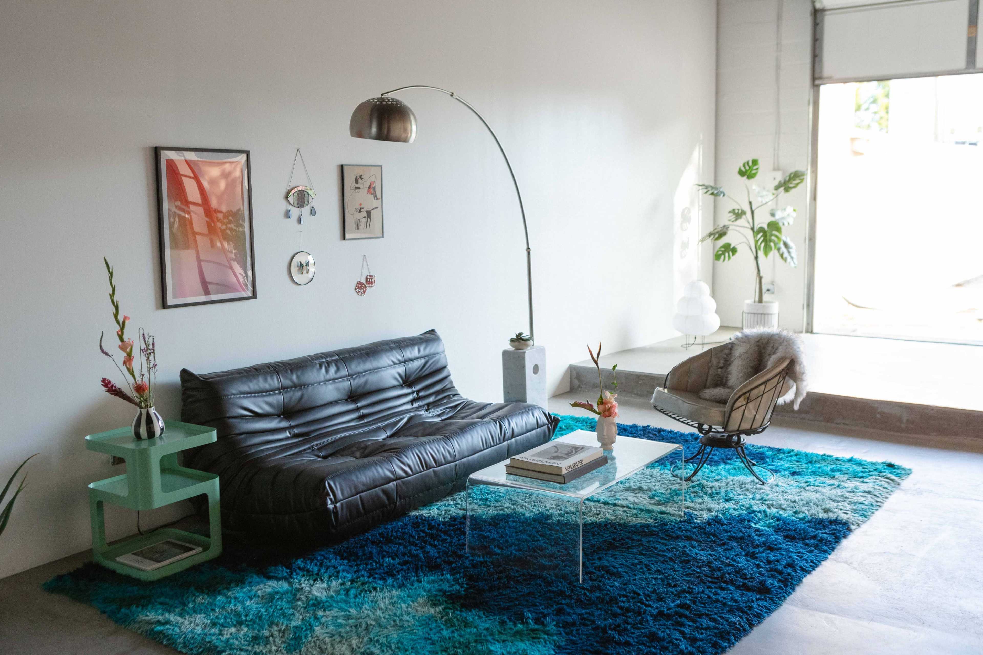 The image shows a modern living room featuring a black leather sofa, a glass coffee table, and an armchair, with colorful rugs and decorative plants enhancing the space.