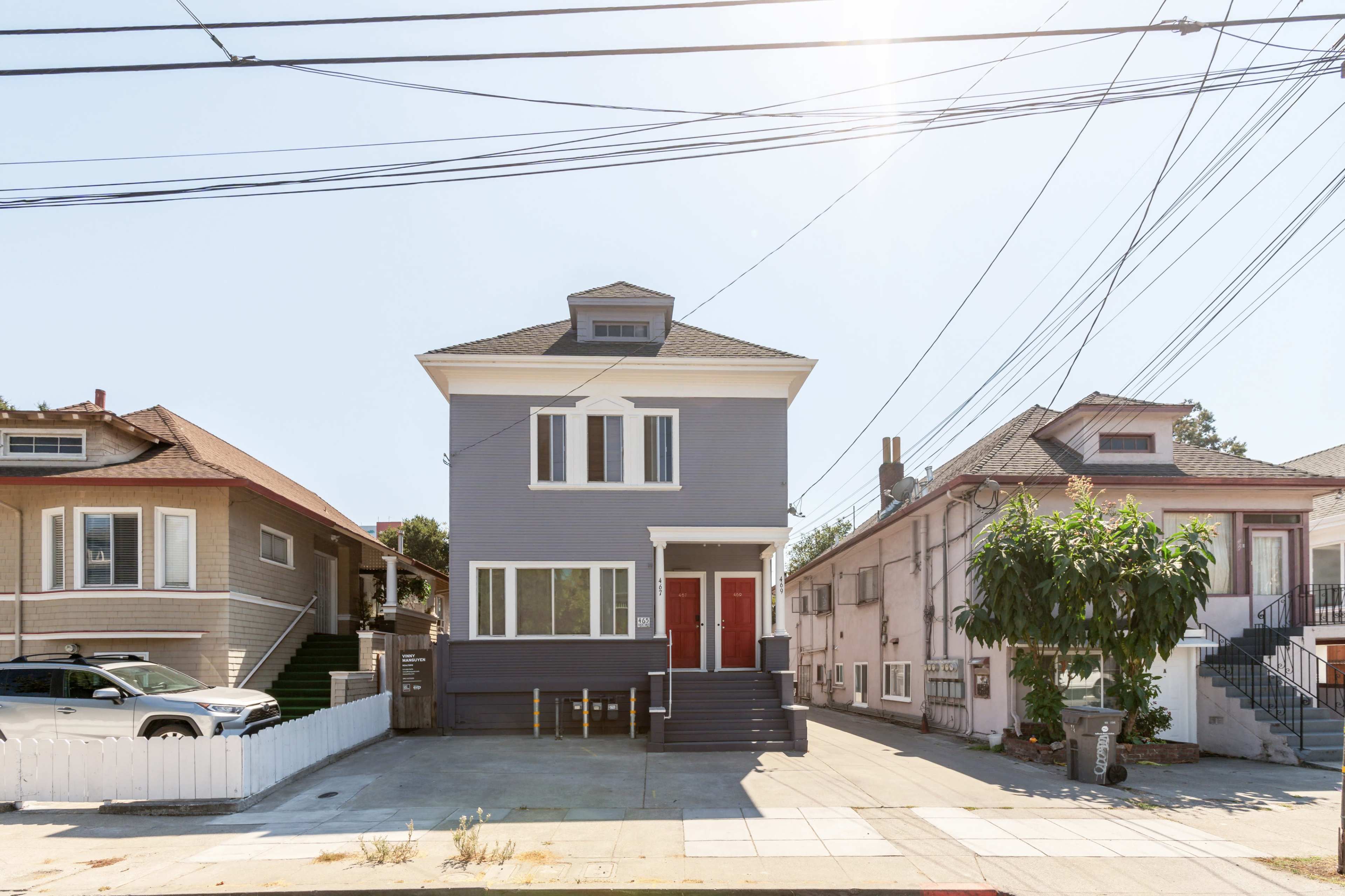A two-story gray house with a red door, flanked by two other residential buildings and surrounded by utility wires on a clear day.
