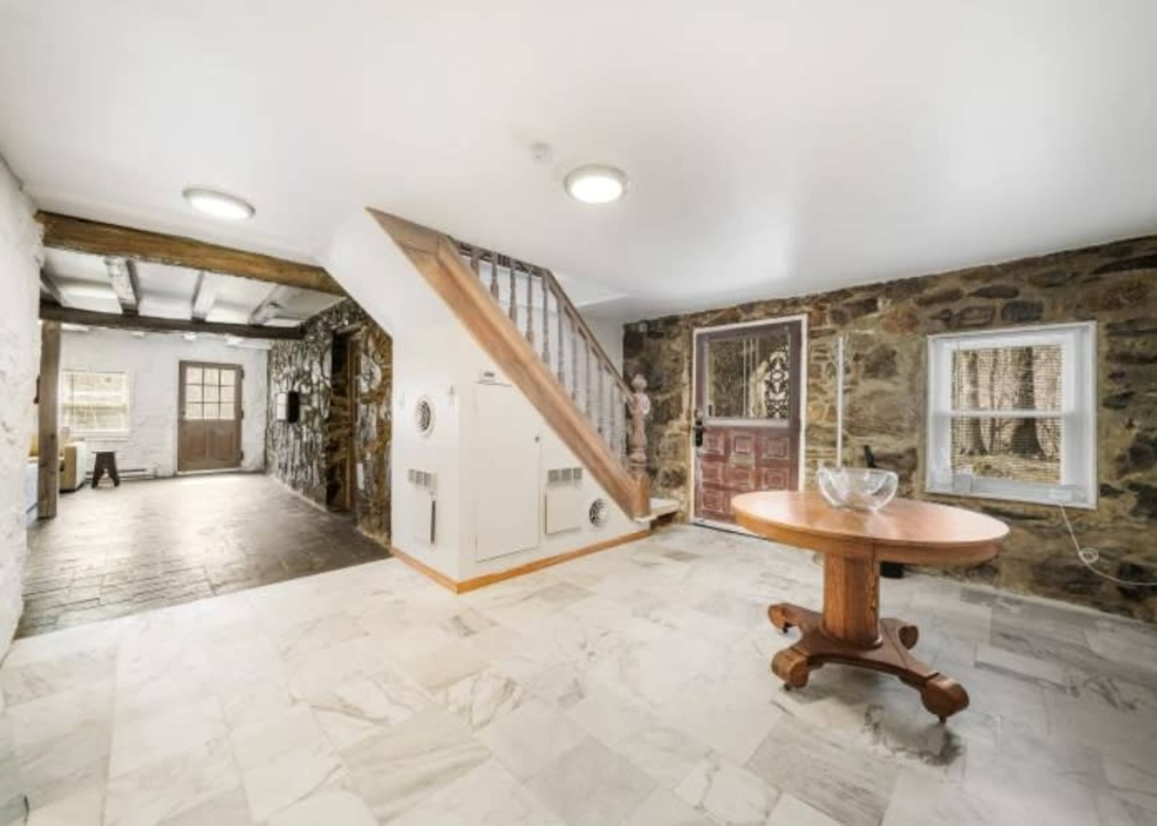 The image shows an interior entryway featuring a wooden table, a staircase leading up, and stone walls with two doorways.