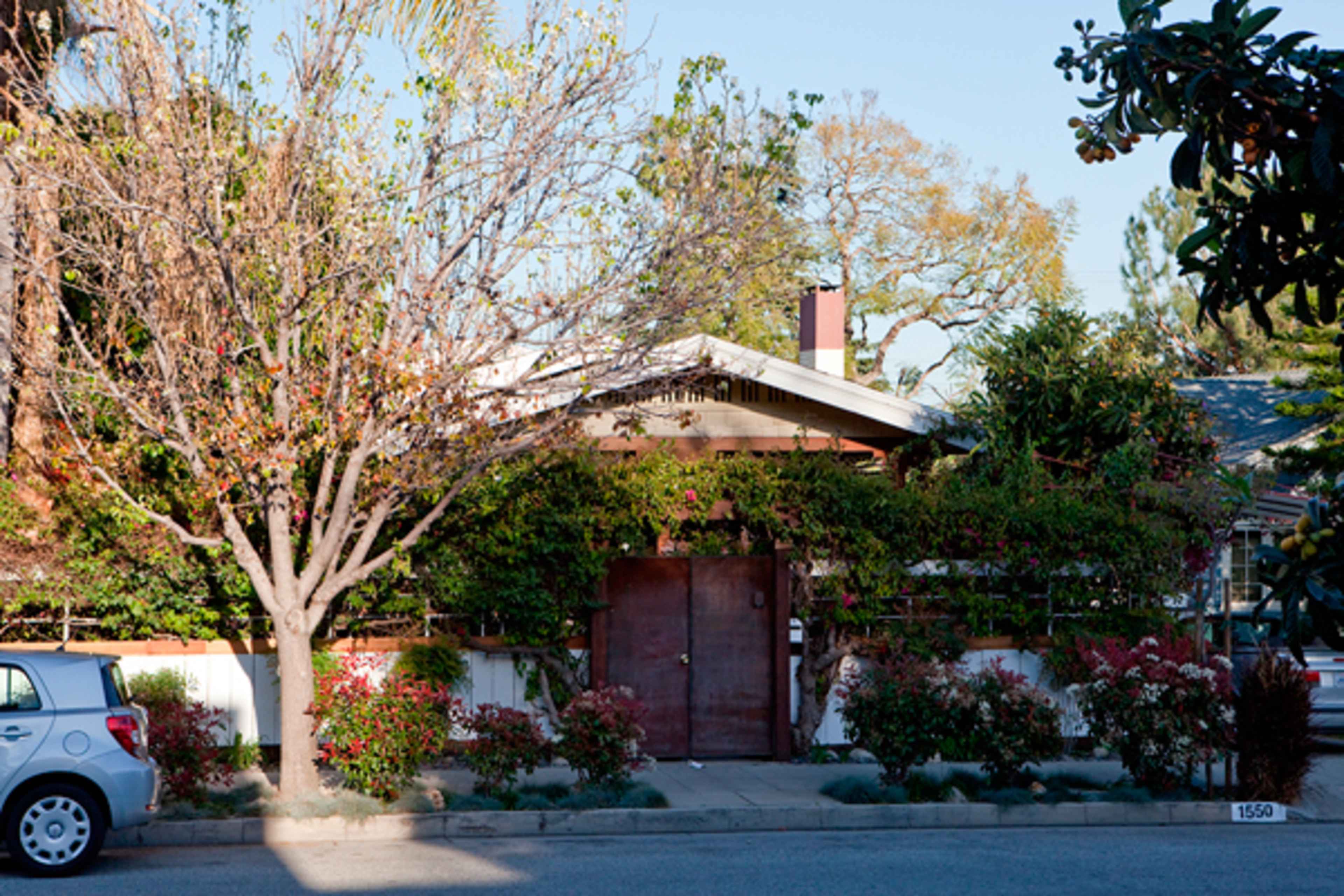 A wooden gate stands at the entrance of a house surrounded by lush greenery and flowering plants, with a parked car nearby on the street.