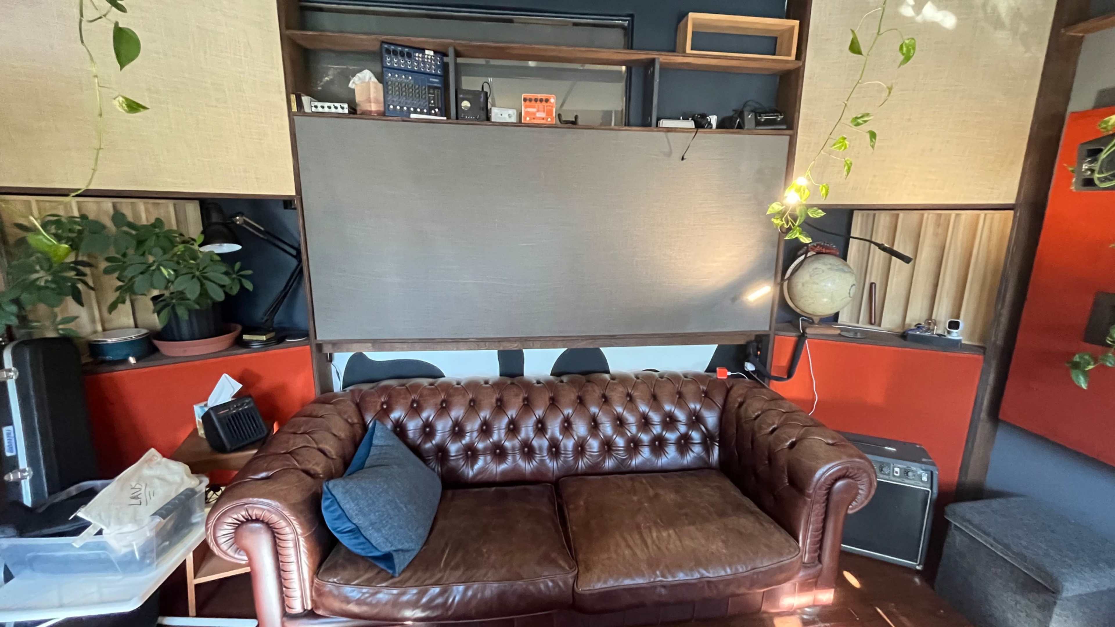 A brown leather Chesterfield sofa is positioned in front of a wall with plants, shelves, and musical equipment in a cozy living room space.