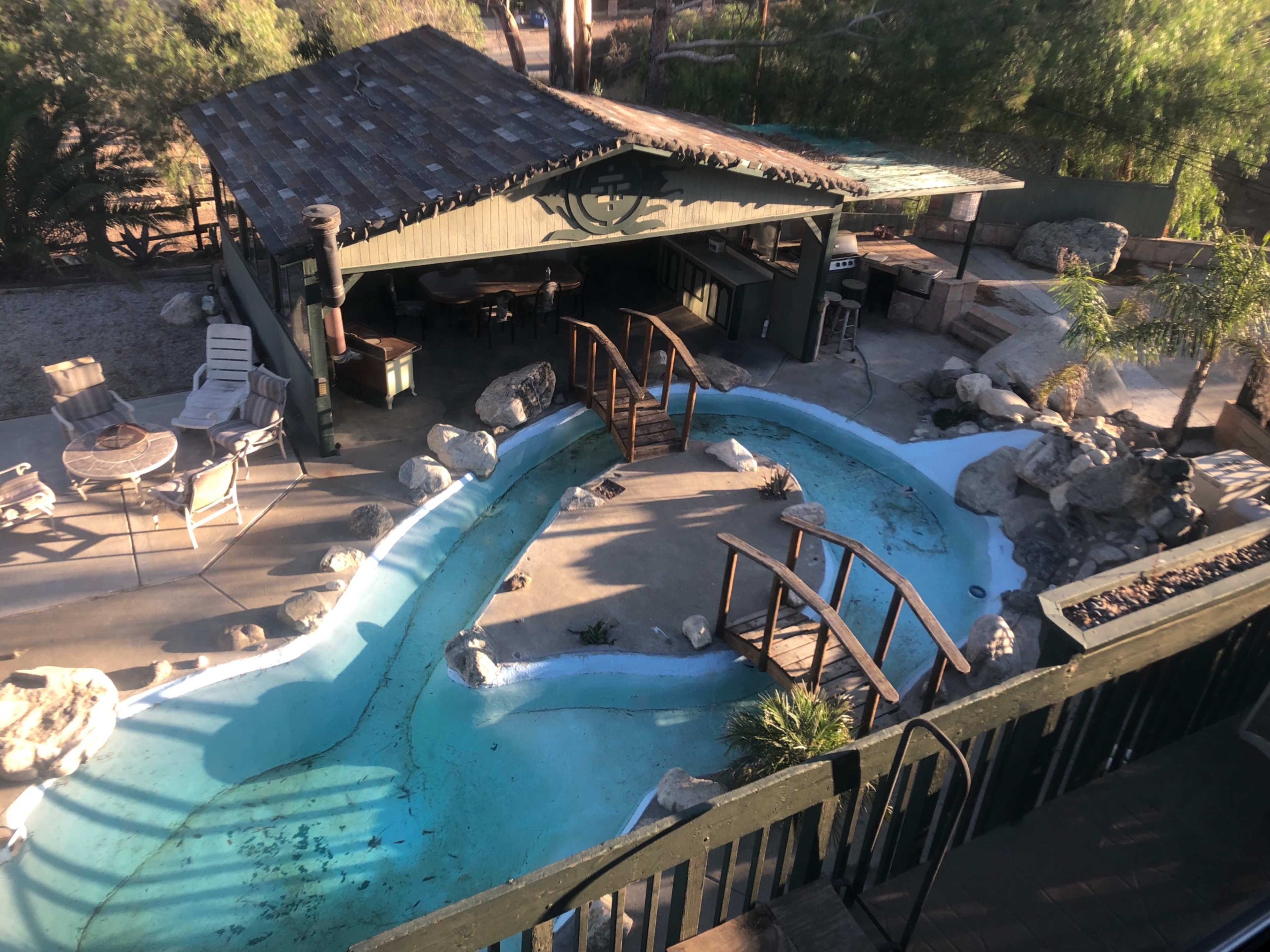 A curved pool area surrounded by rocks and a wooden bridge, with a shaded seating area nearby.