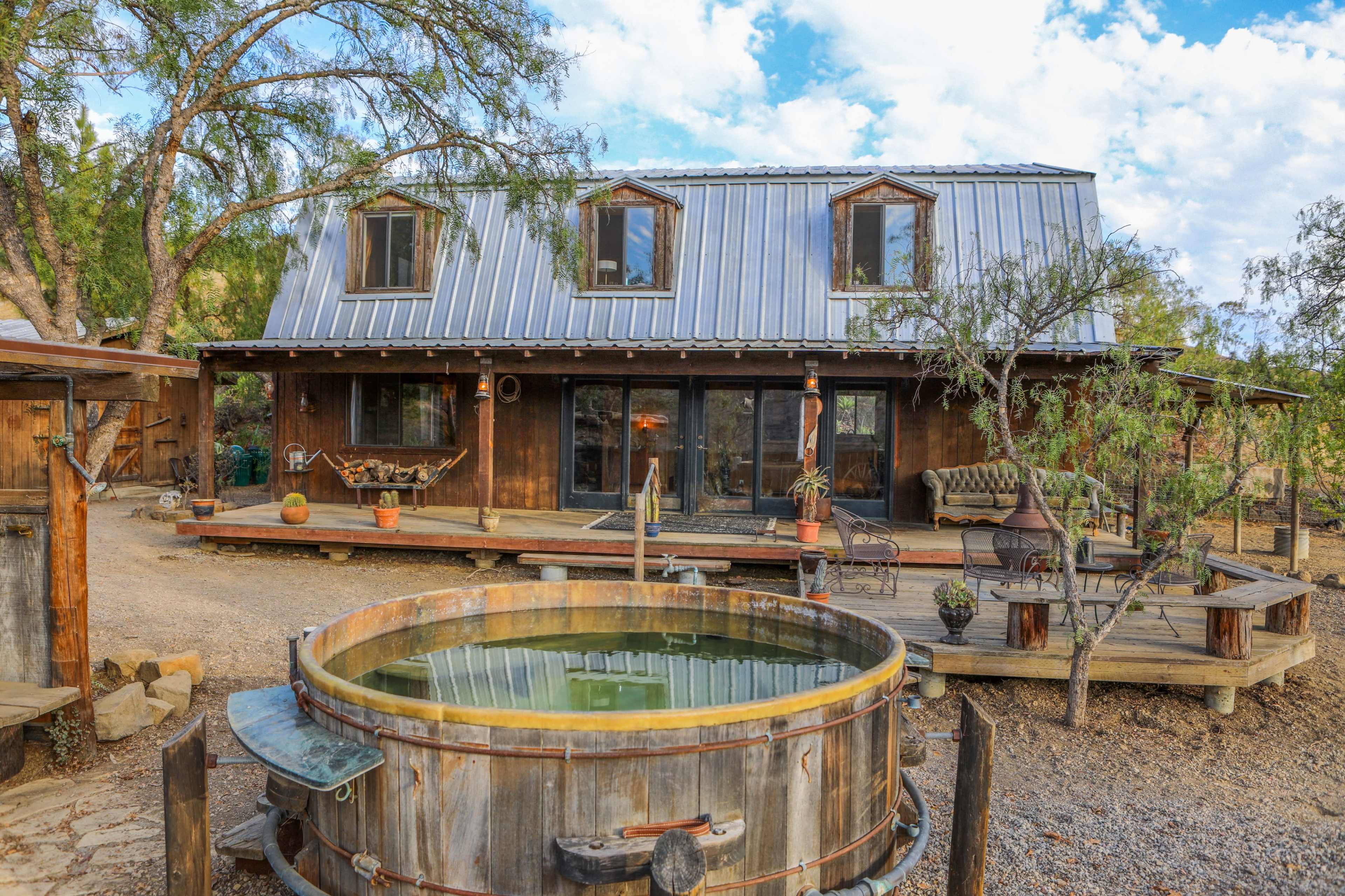 A rustic wooden house with a metal roof, surrounded by various trees, features a large wooden hot tub on a deck in the foreground.