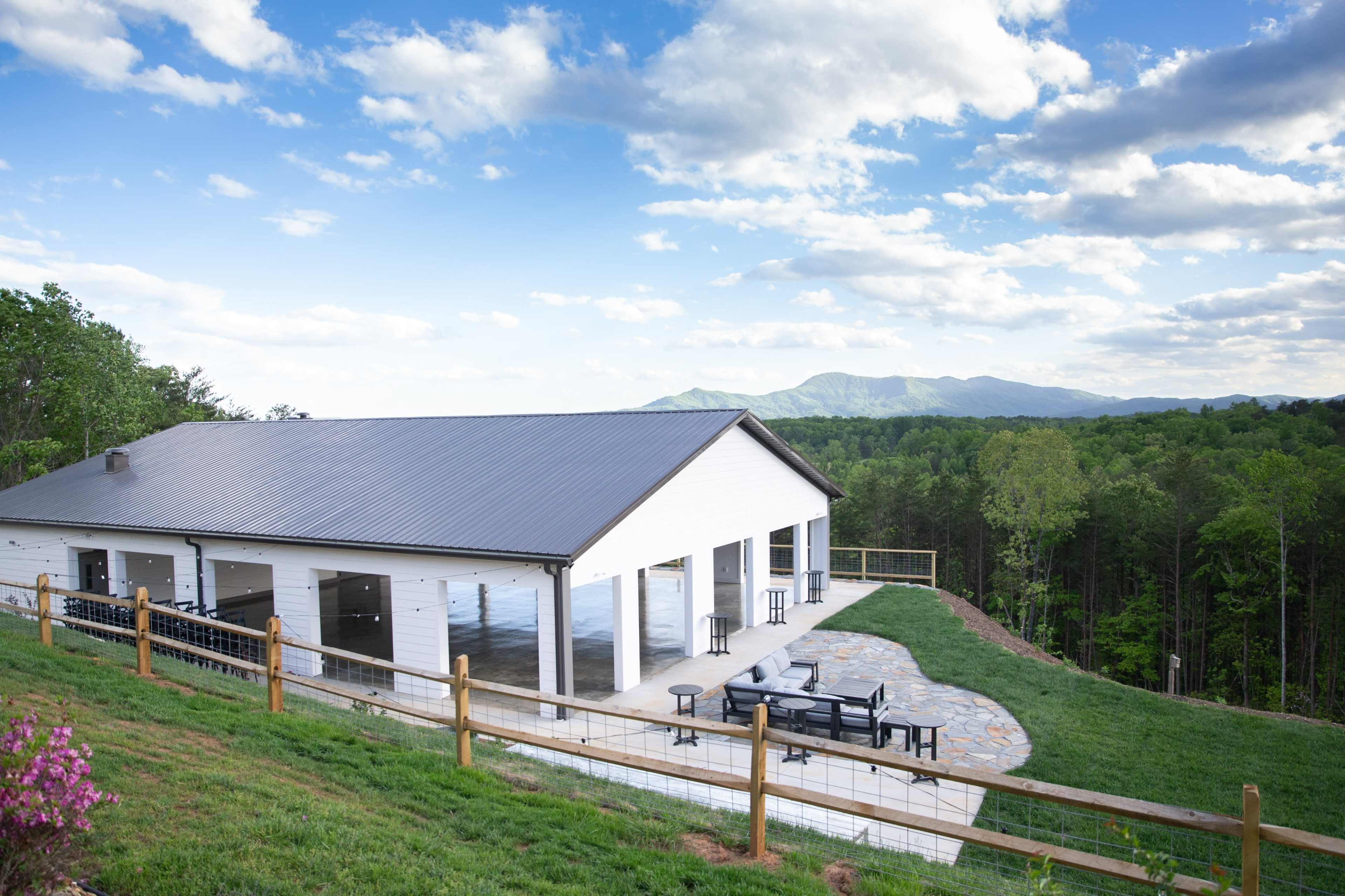 A white building with a metal roof is situated on a hillside, overlooking a green landscape and distant mountains under a partly cloudy sky.