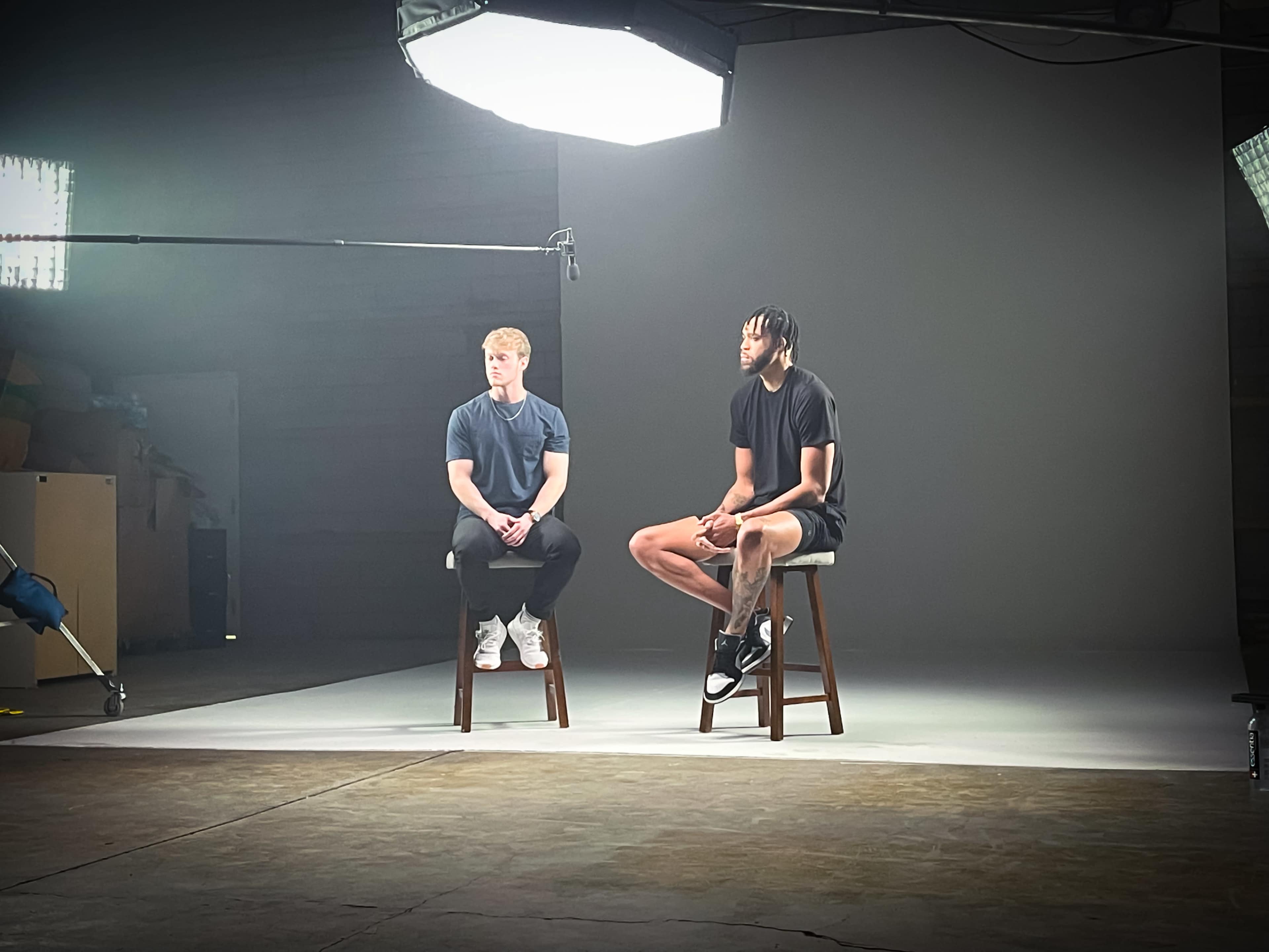 Two men sit on stools in a studio setting, with bright lights and a gray backdrop.
