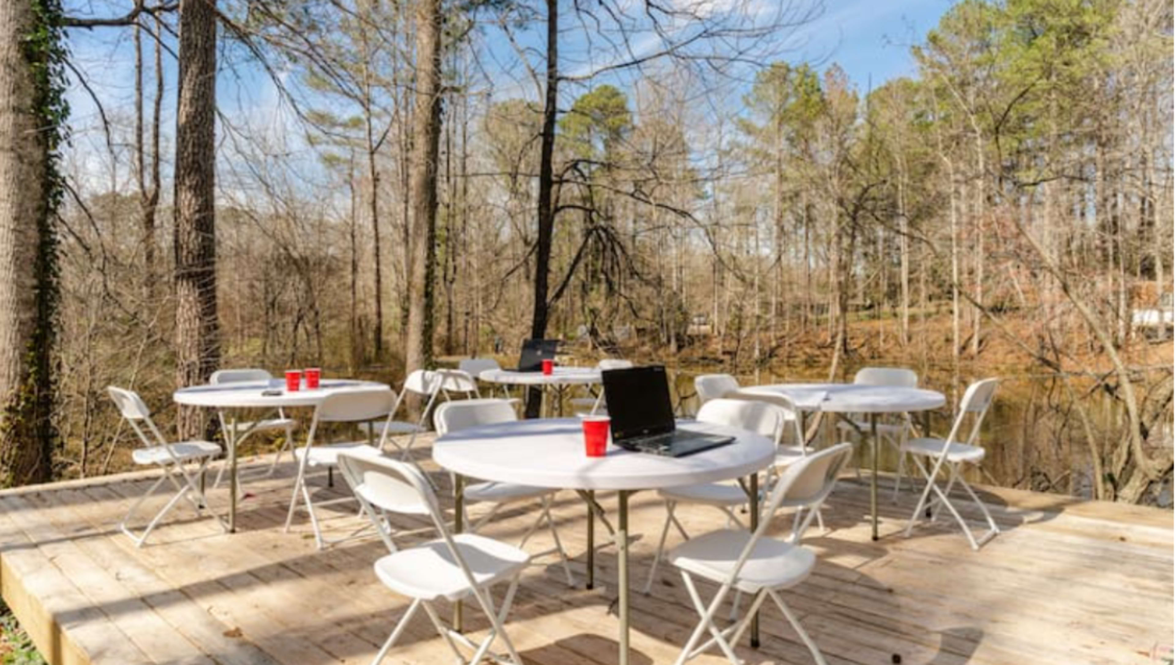 A wooden deck beside a pond is furnished with several white folding tables and chairs, along with a laptop and red cups.