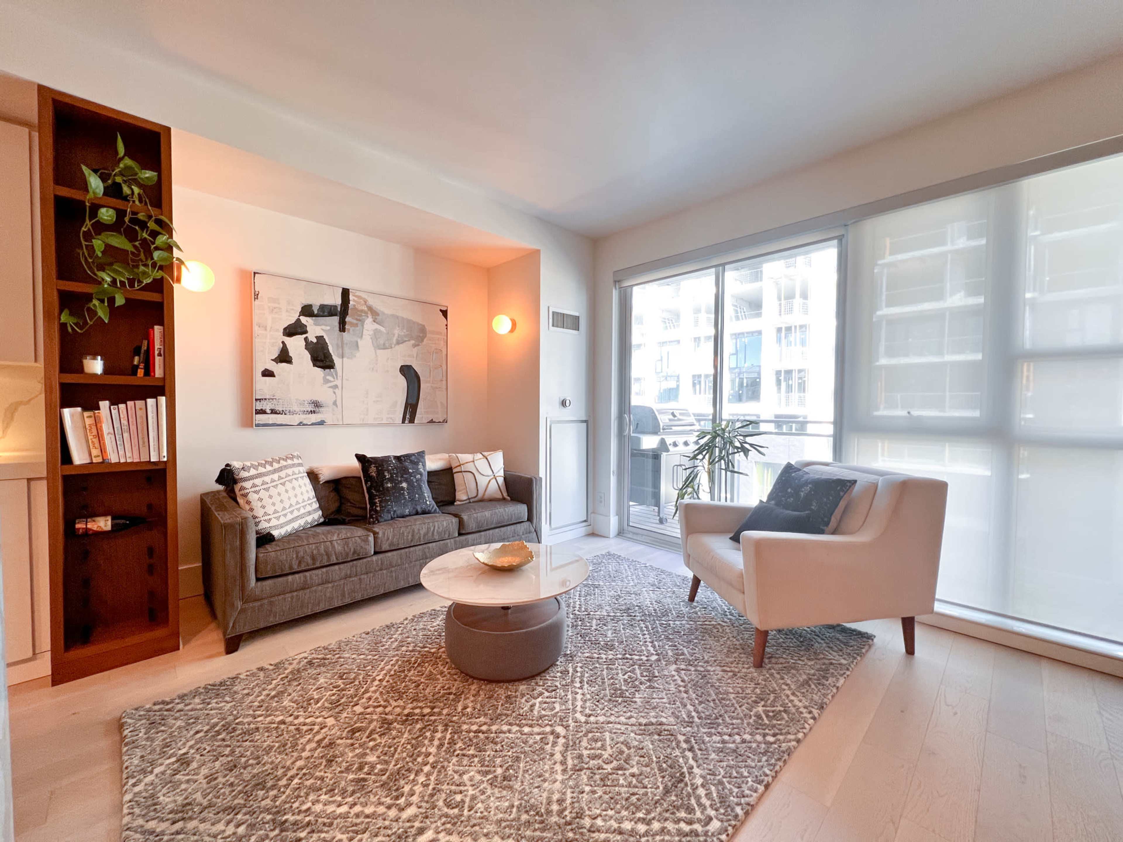 A modern living room featuring a gray sofa, a light-colored armchair, a round coffee table, and large windows allowing natural light.