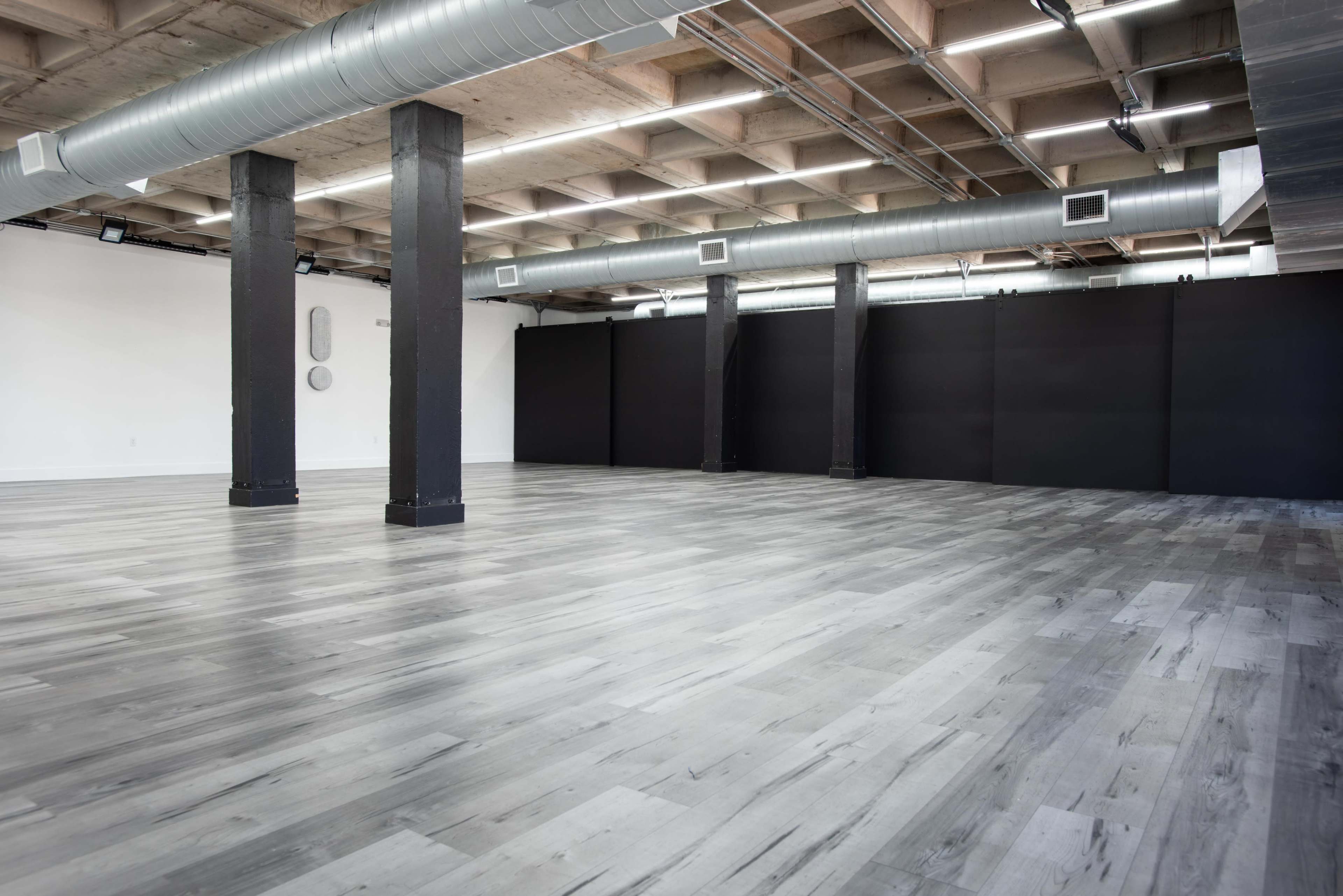 A spacious, empty room with polished gray wooden floors and black walls, supported by dark columns.