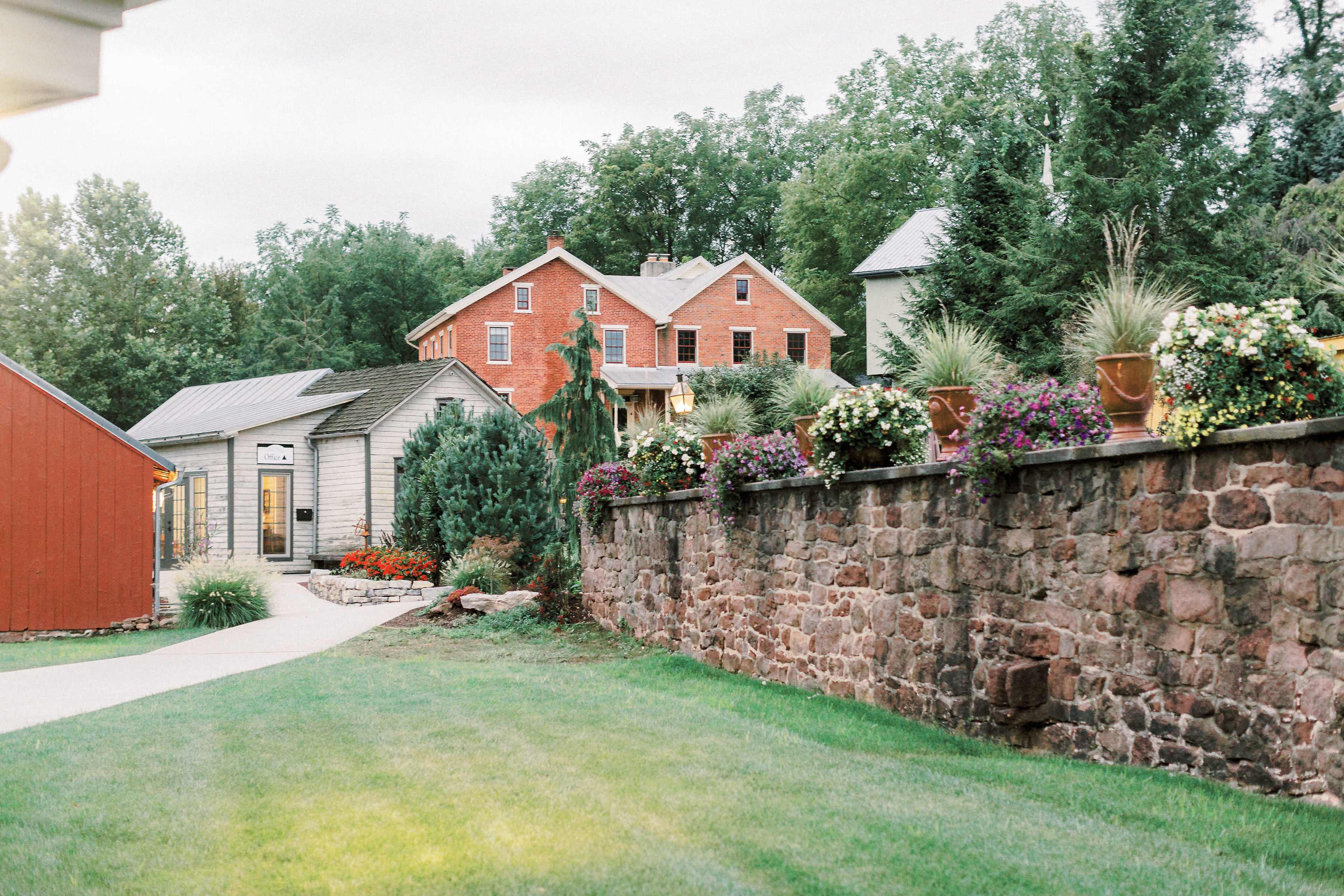 A landscaped garden with colorful flower pots is framed by a stone wall, leading to a red brick house and a nearby gray building in the background.