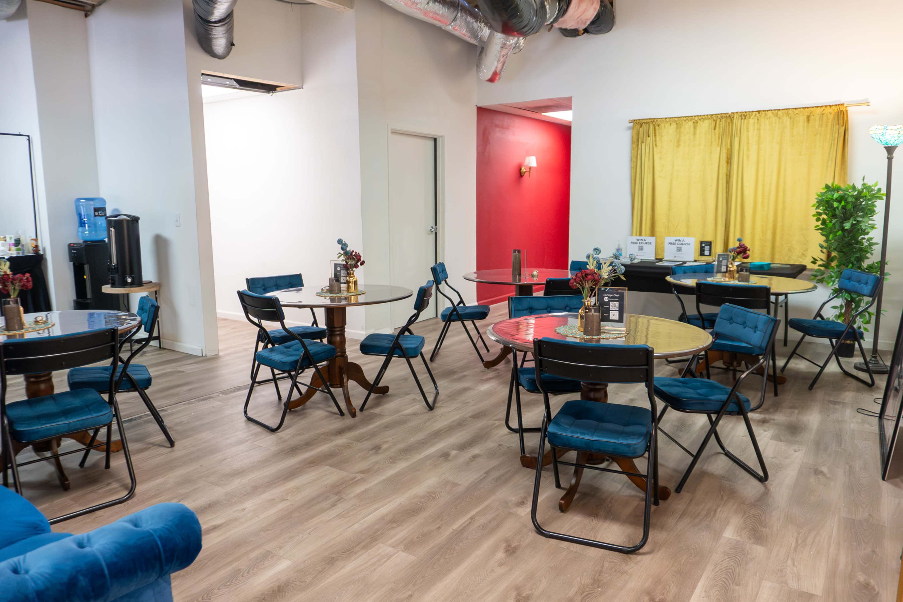 The image shows a bright, modern room with several round tables surrounded by blue chairs, a water dispenser, and a yellow curtain in the background.
