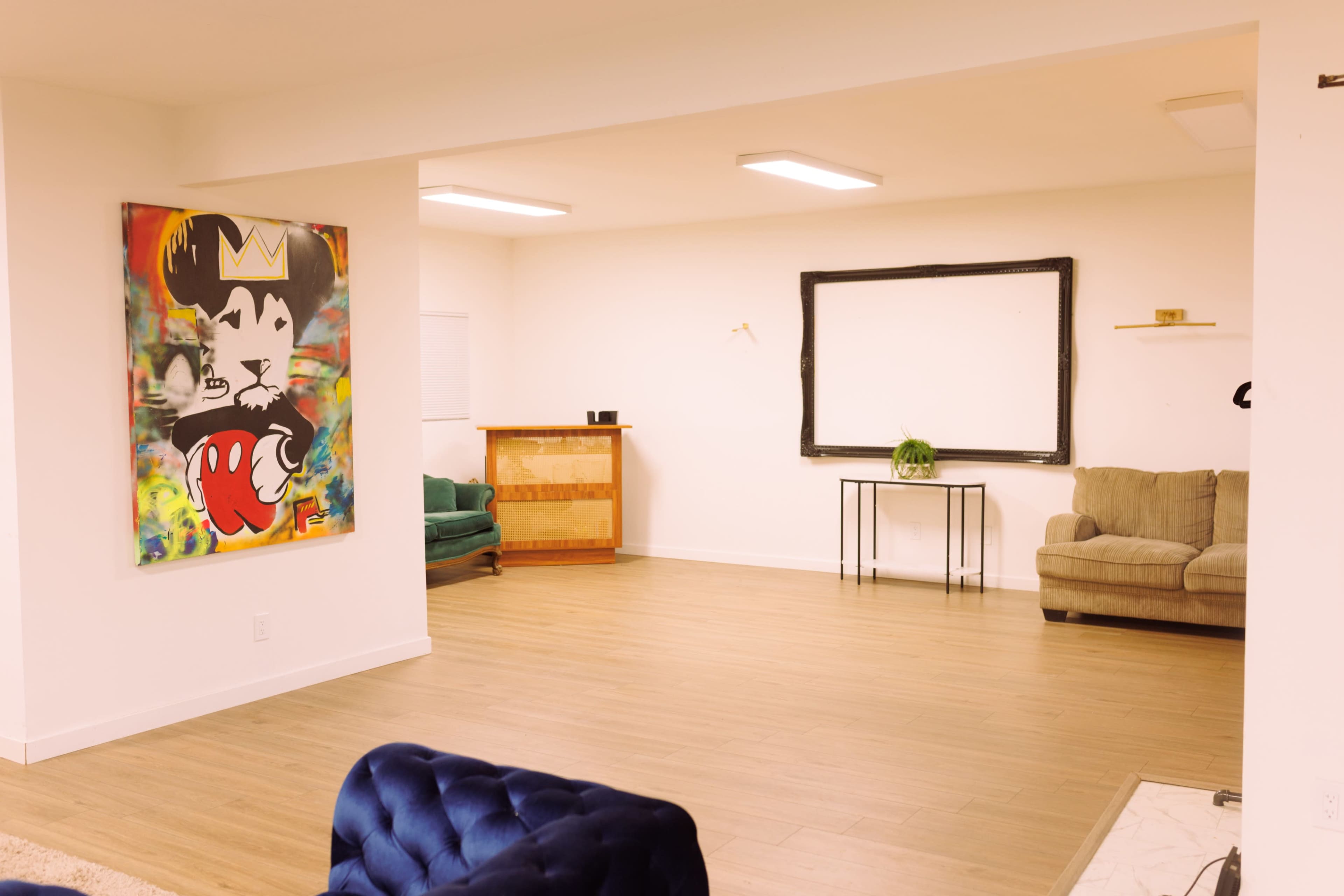 The image shows a spacious, sparsely furnished room with light wood flooring, a large artwork on the wall, and a few pieces of furniture including green and gray sofas.