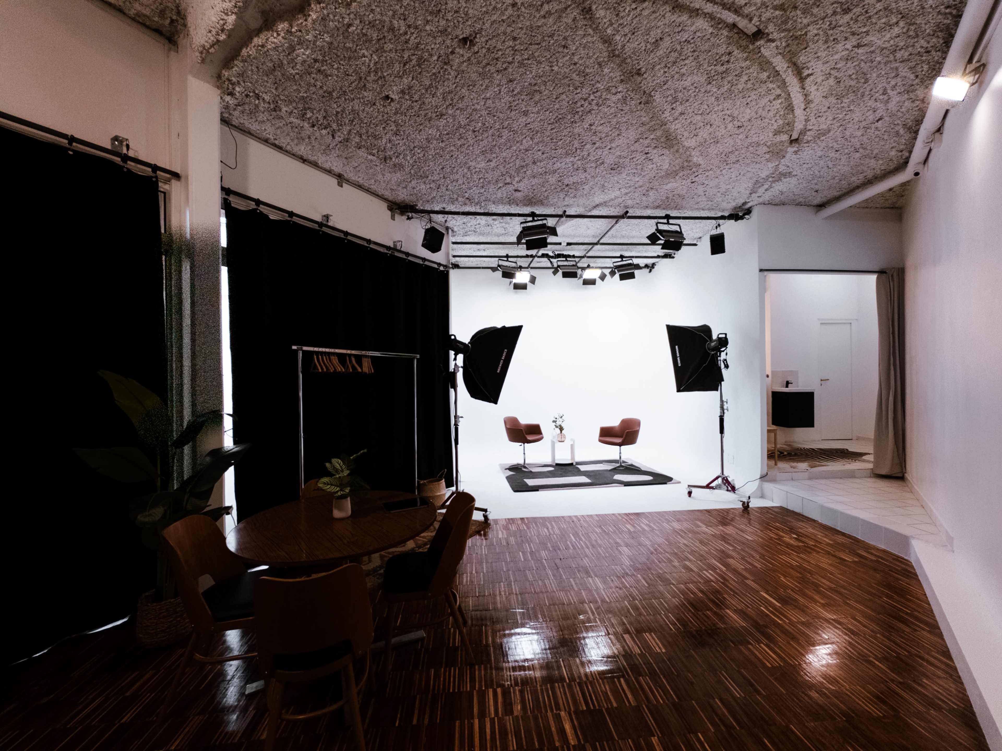 The image shows a well-lit studio with two chairs on a set, complemented by large softbox lights and a dining area with a wooden table and chairs.