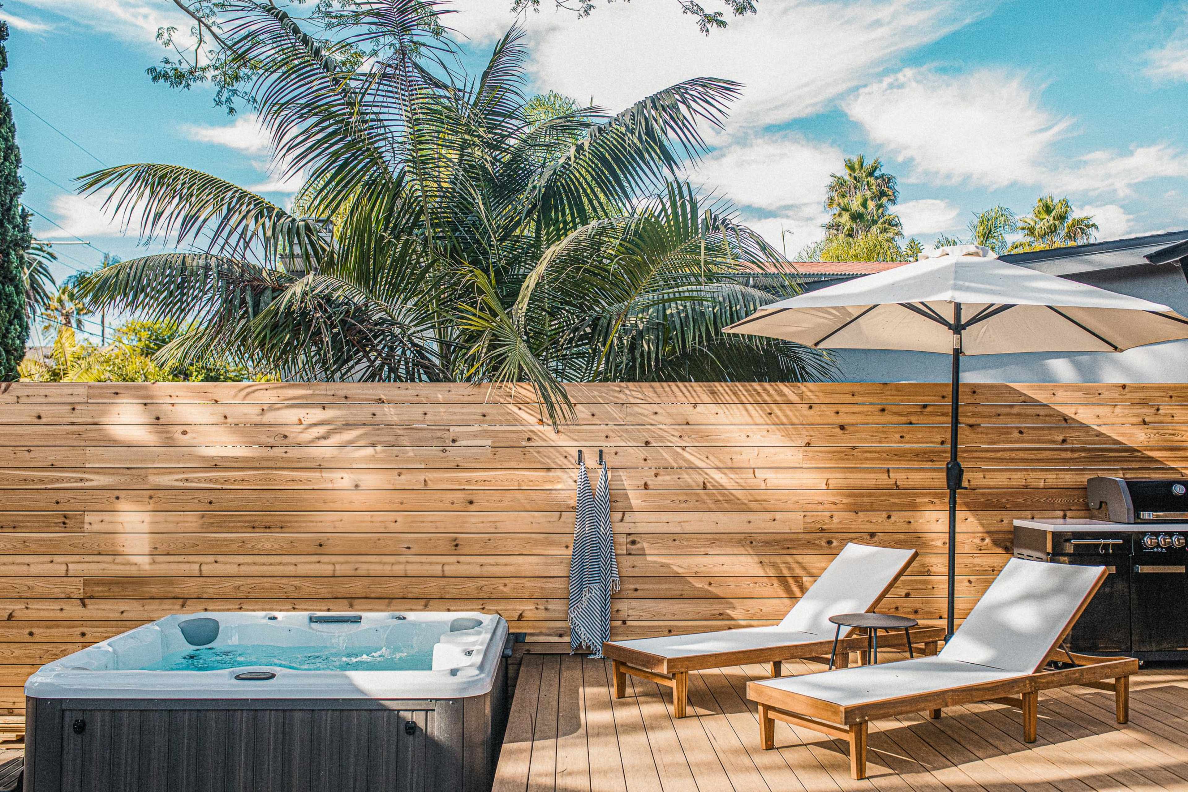 A backyard featuring a hot tub, two lounge chairs, an umbrella, and a wooden fence surrounded by palm trees.