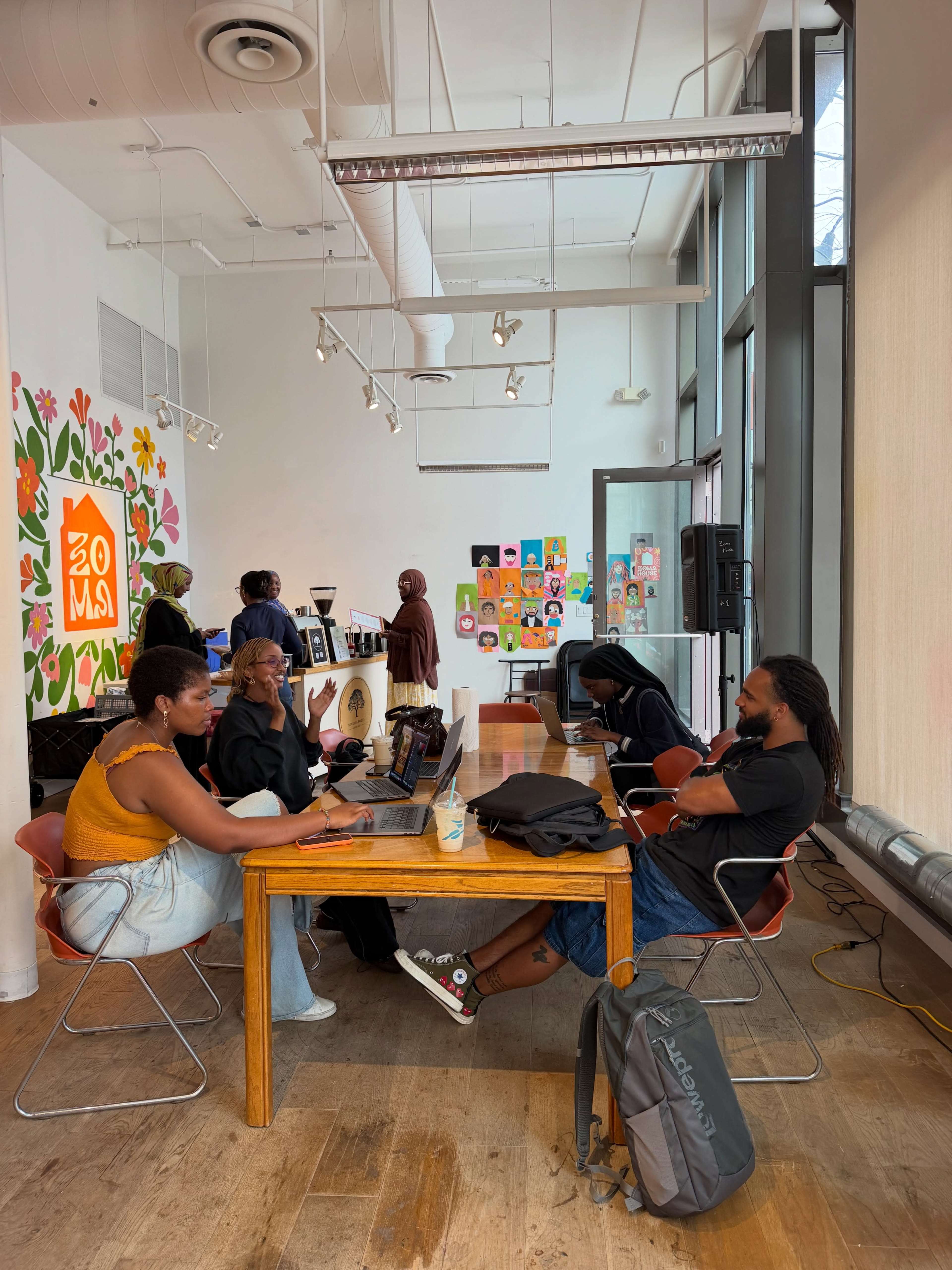 A group of people are seated at a wooden table in a spacious room with art on the walls, engaged in conversation and working on laptops.