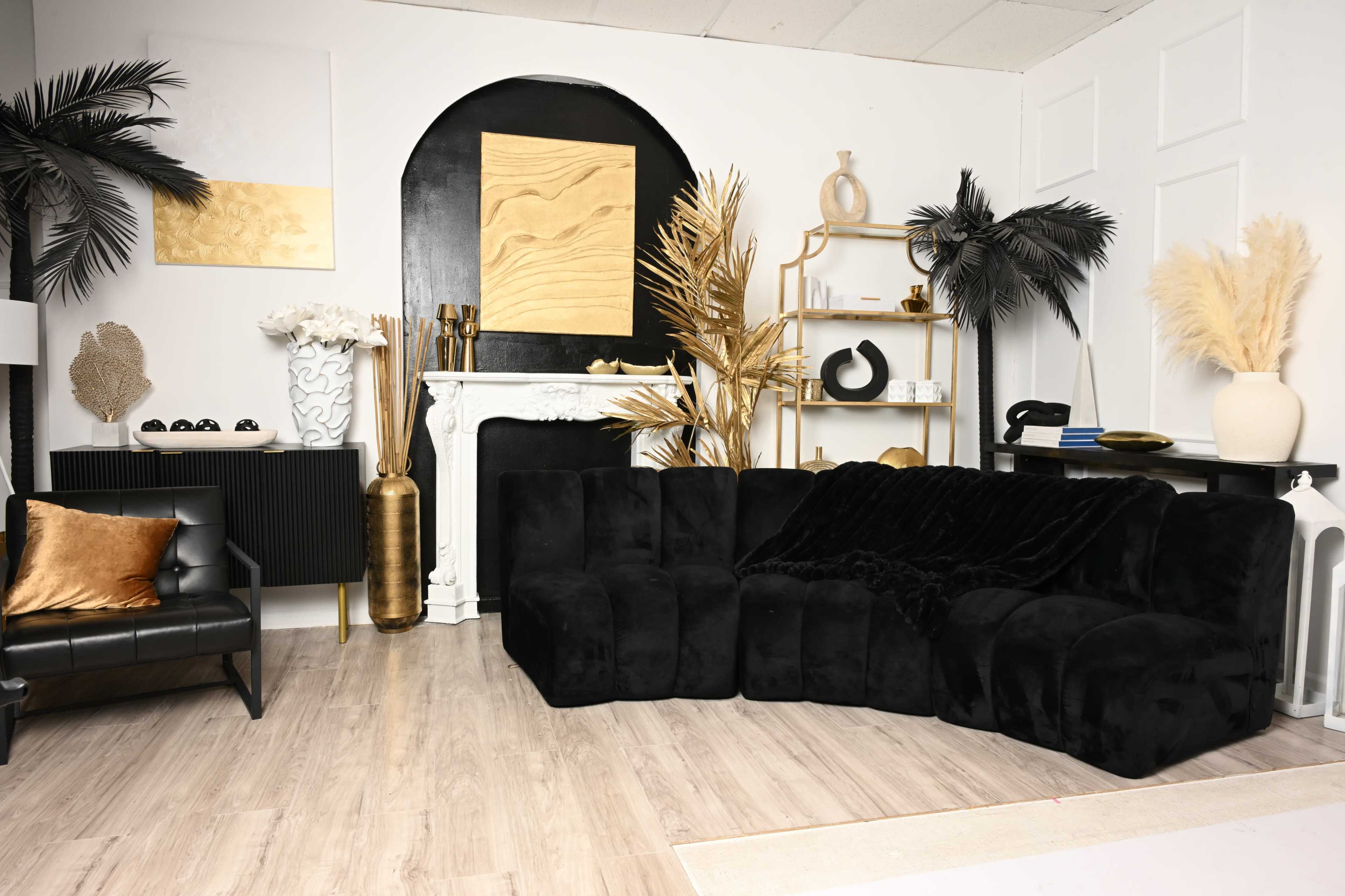 The image shows a stylish interior space featuring a large black sectional sofa, a black leather chair, decorative plants, gold accents, and various art pieces on the walls.