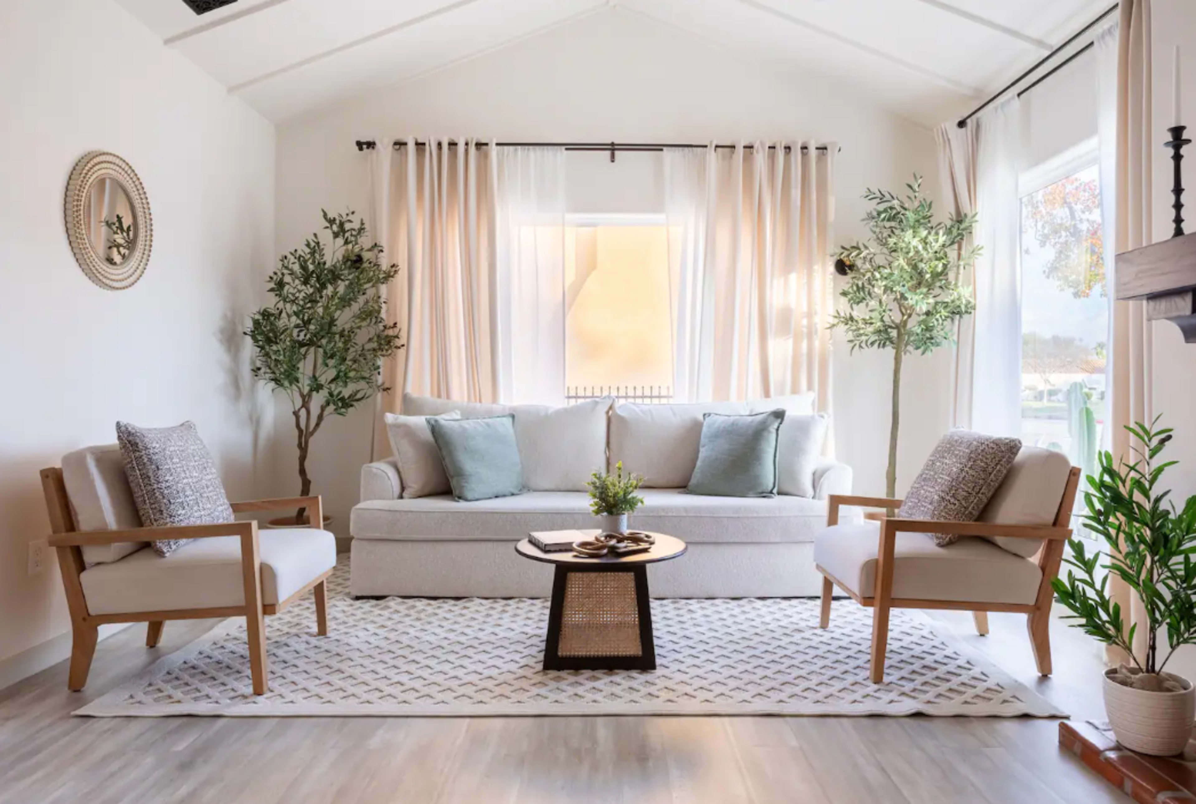 A light and airy living room features a sofa, two armchairs, a round coffee table, and large windows dressed with sheer curtains.