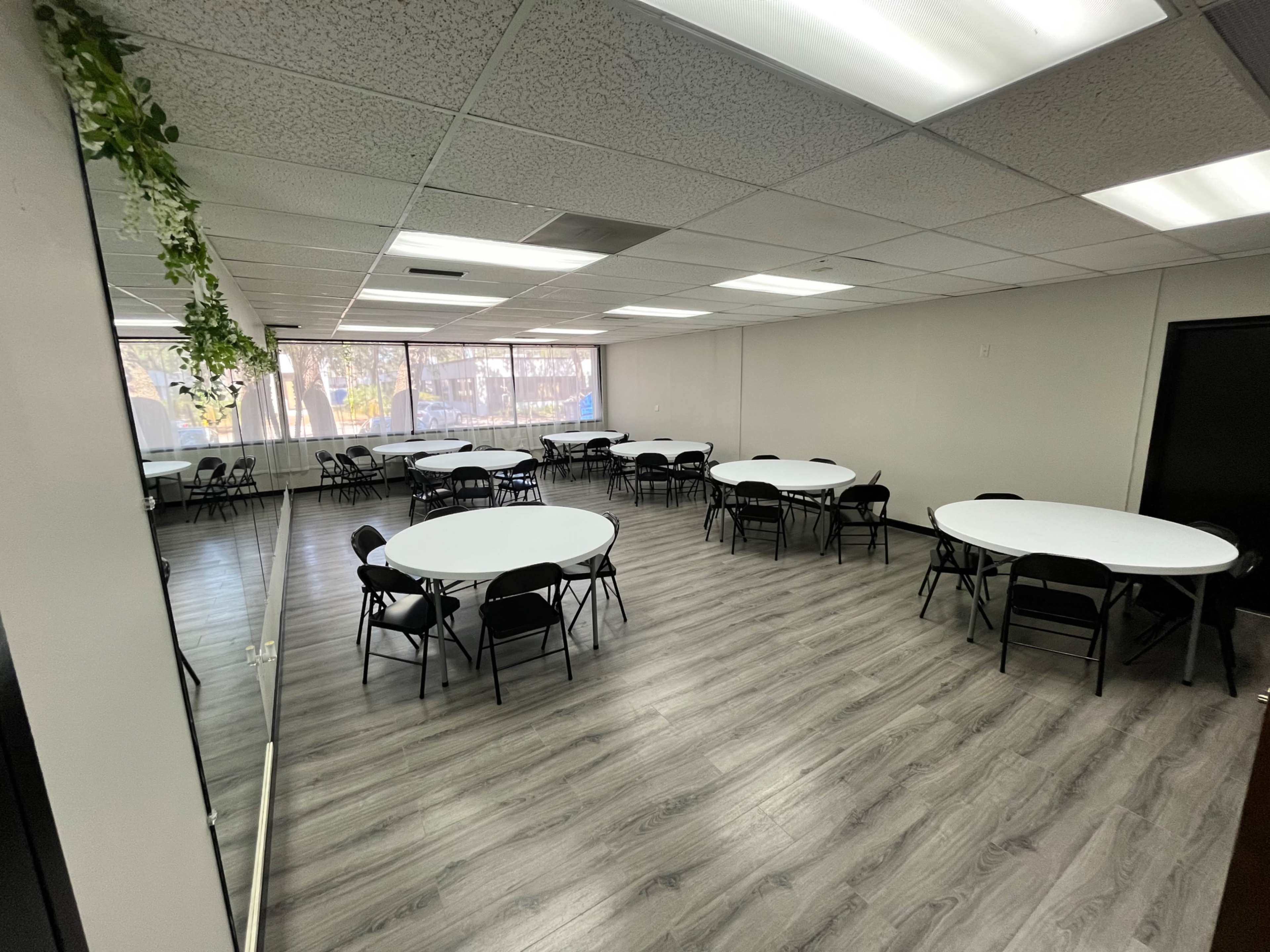 The image shows a spacious room with multiple round tables and black chairs arranged for an event, featuring large windows and a mirror on one wall.