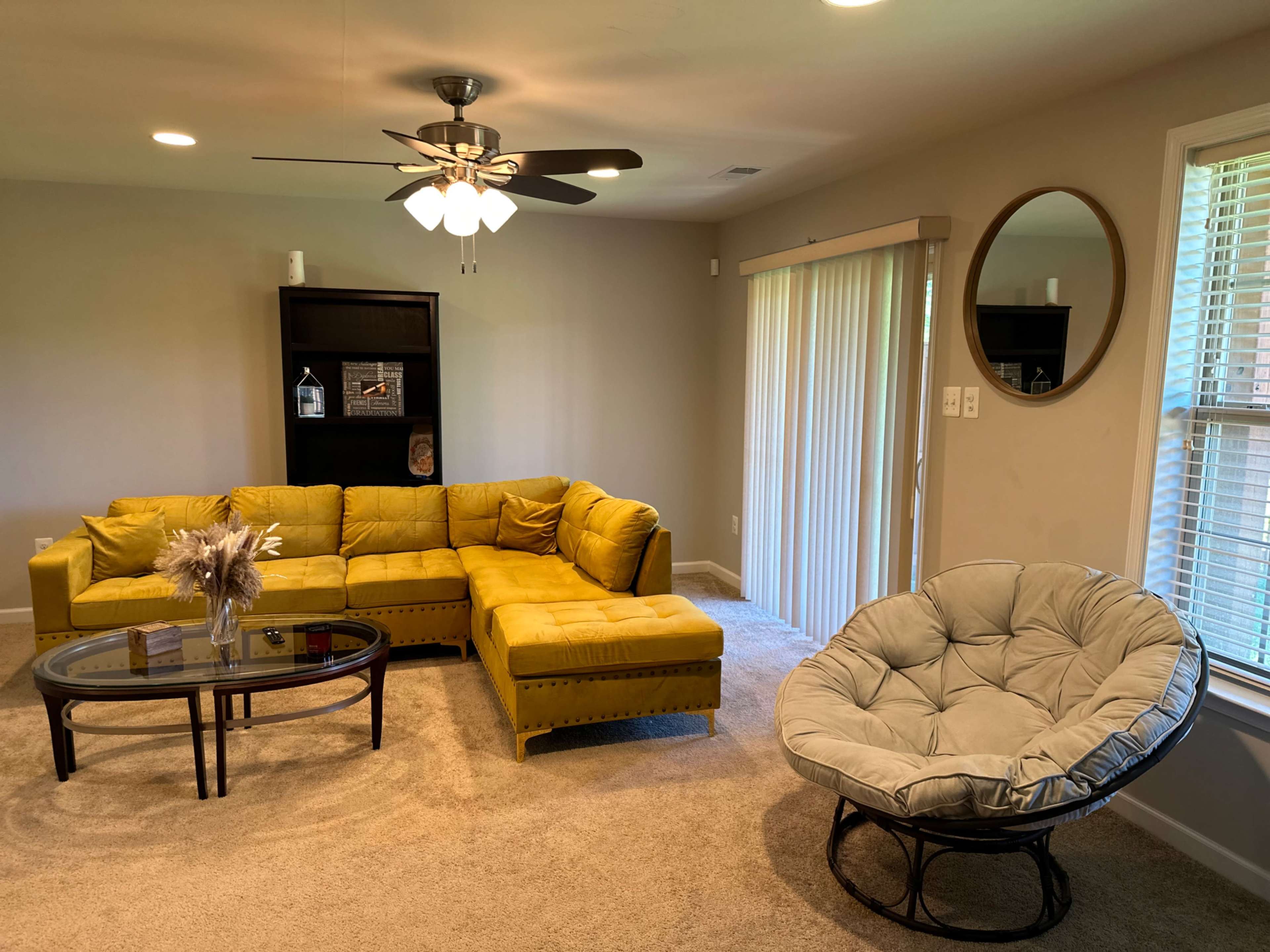 A living room with a yellow sectional sofa, a glass coffee table, a round mirror, and a black chair.