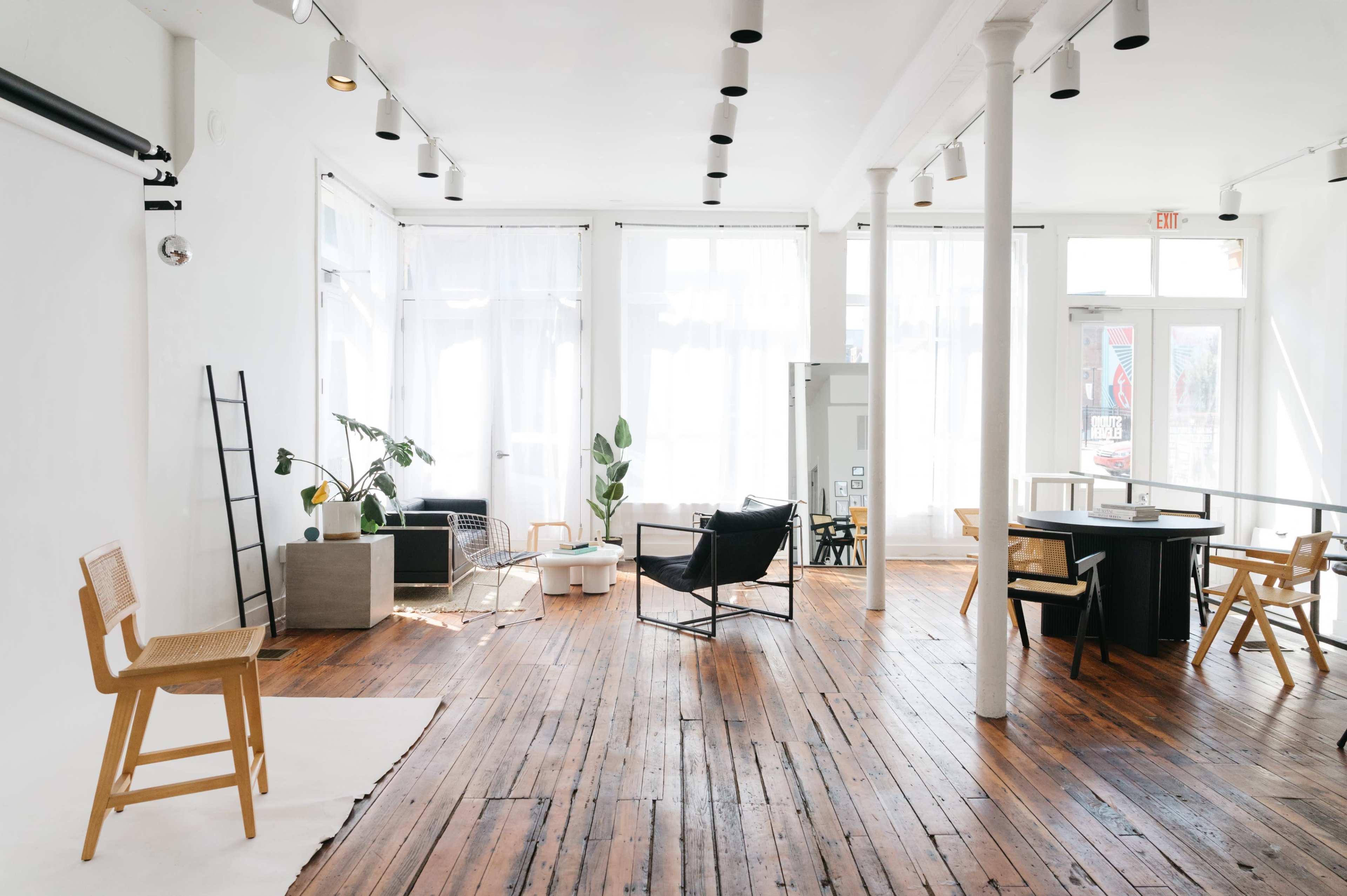 The image shows a bright, modern open space with wooden floors, featuring furniture like chairs and tables, large windows, and plants.