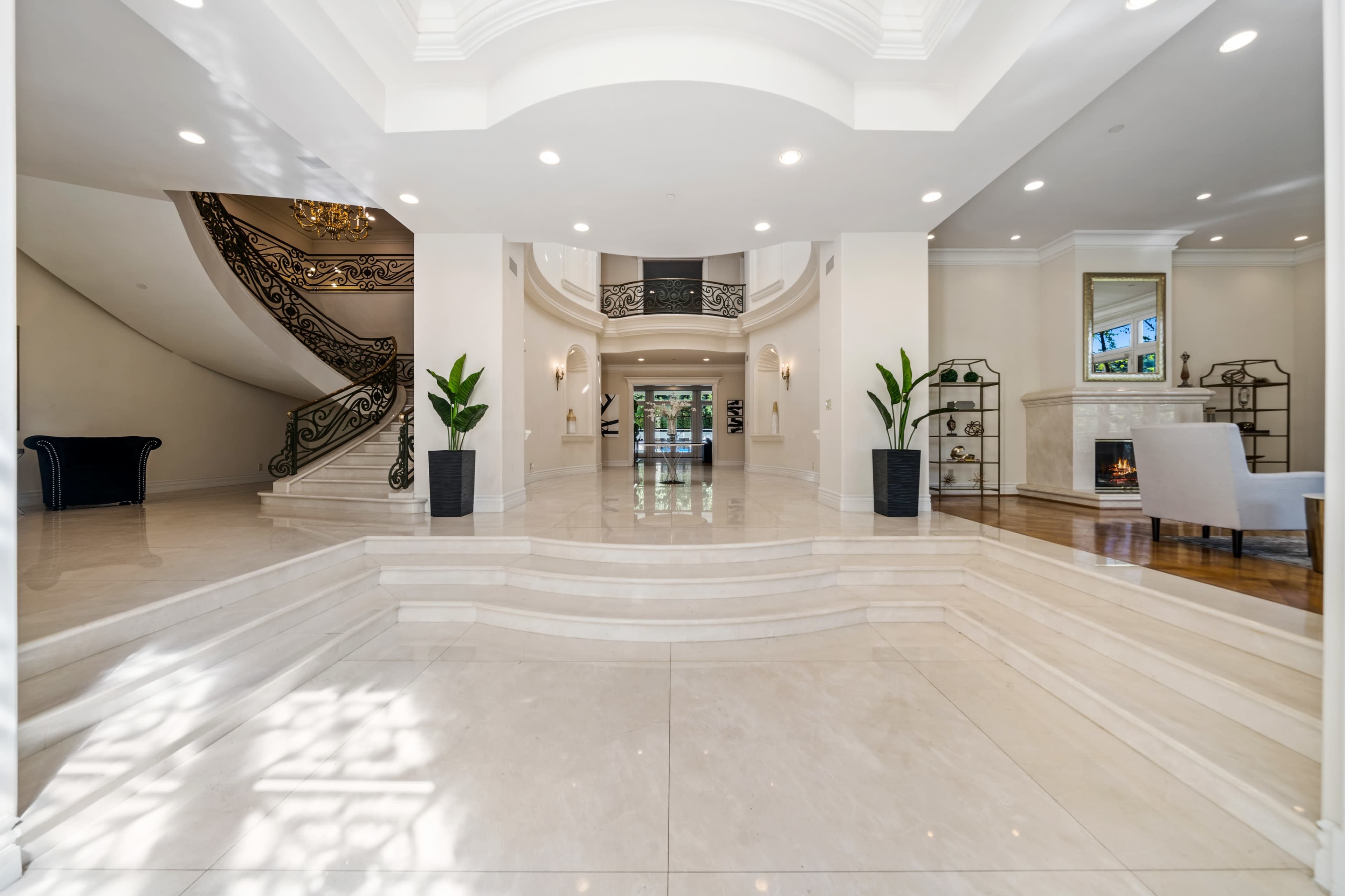 The image shows a spacious foyer with a sweeping staircase, elegant furnishings, and large windows allowing natural light to fill the area.