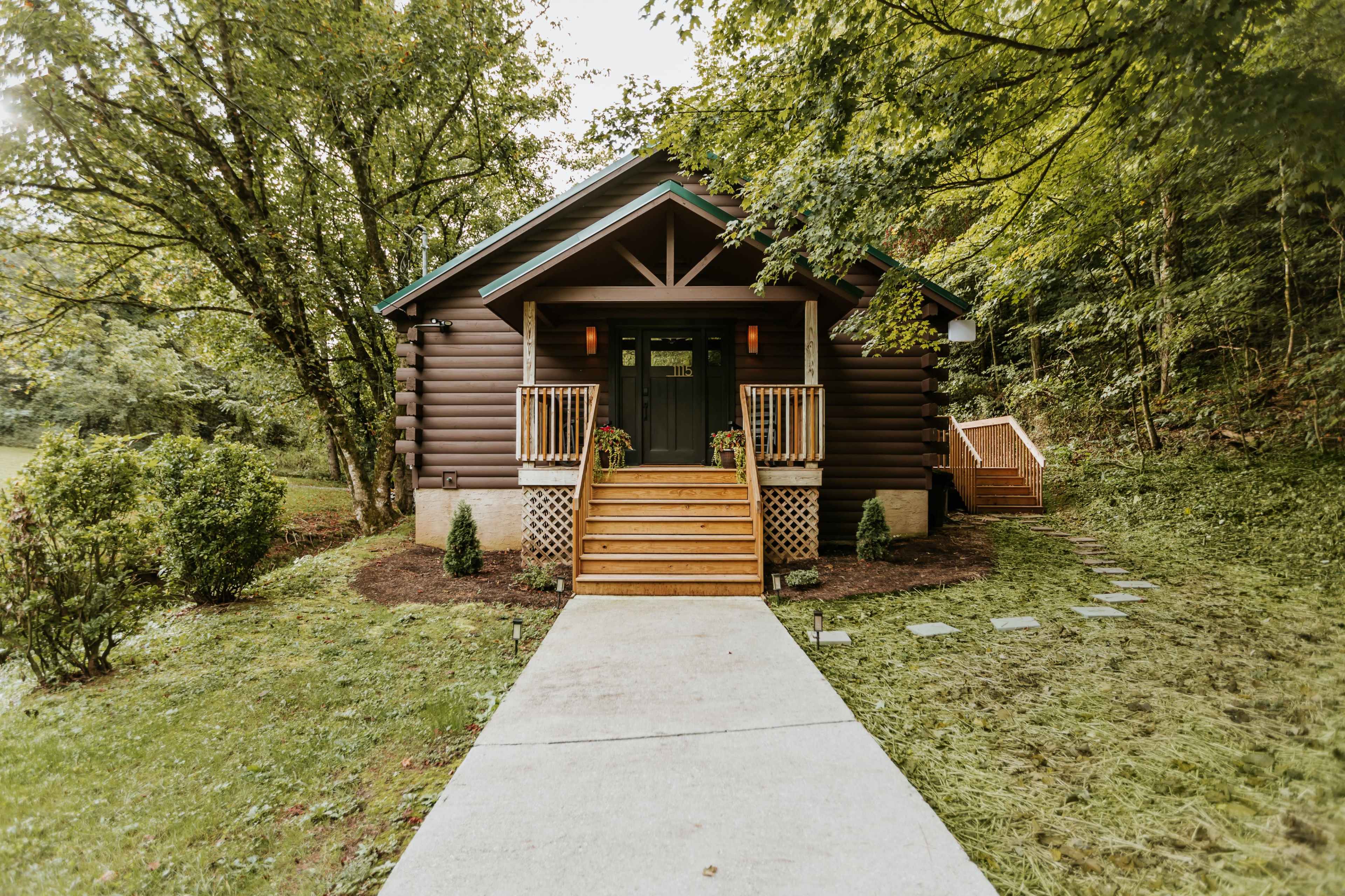 A log cabin with a front porch and wooden steps is set among greenery, leading to a path.