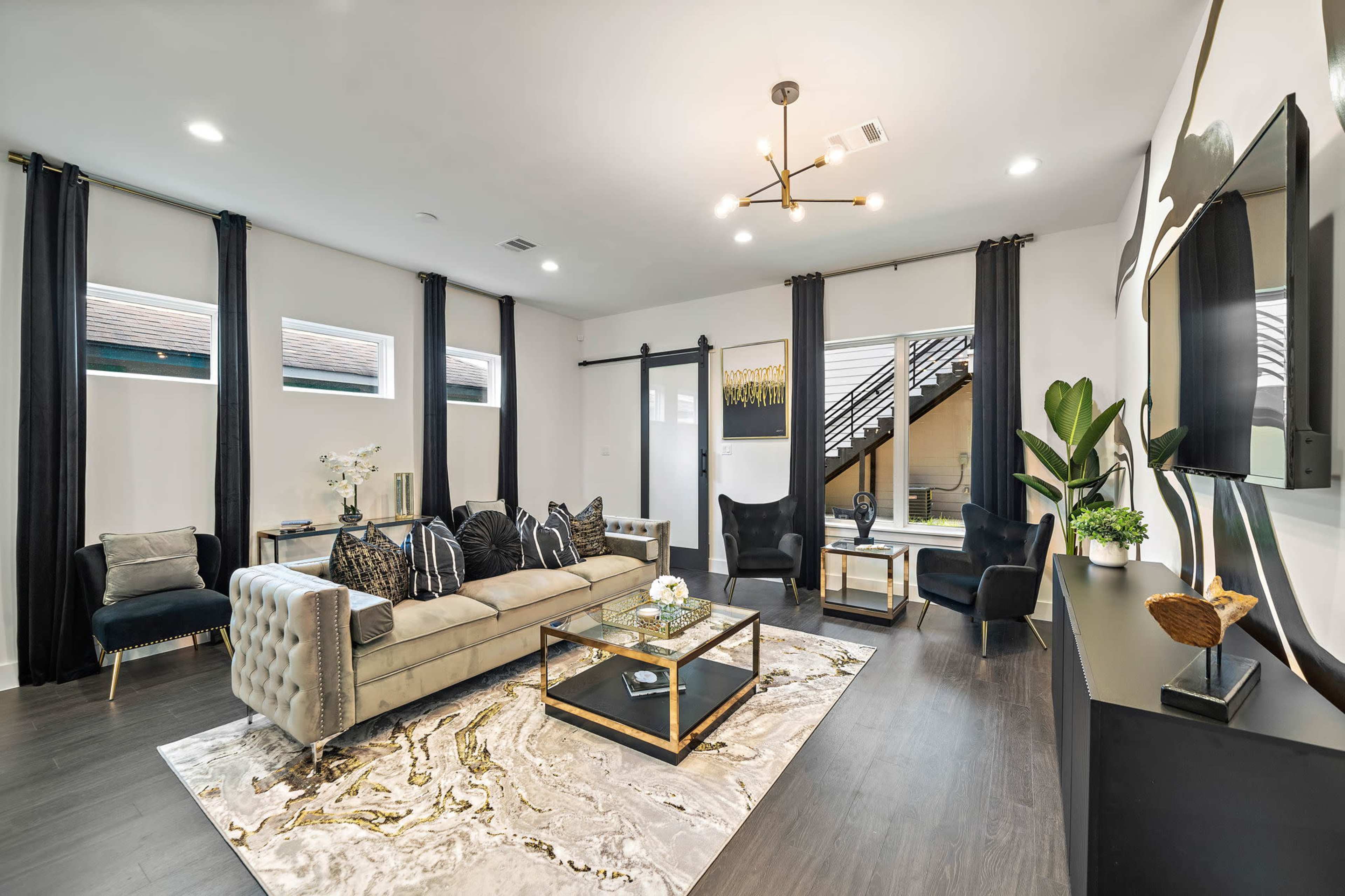The living room features a light-colored sectional sofa, a glass coffee table on a patterned rug, and dark curtains framing large windows.