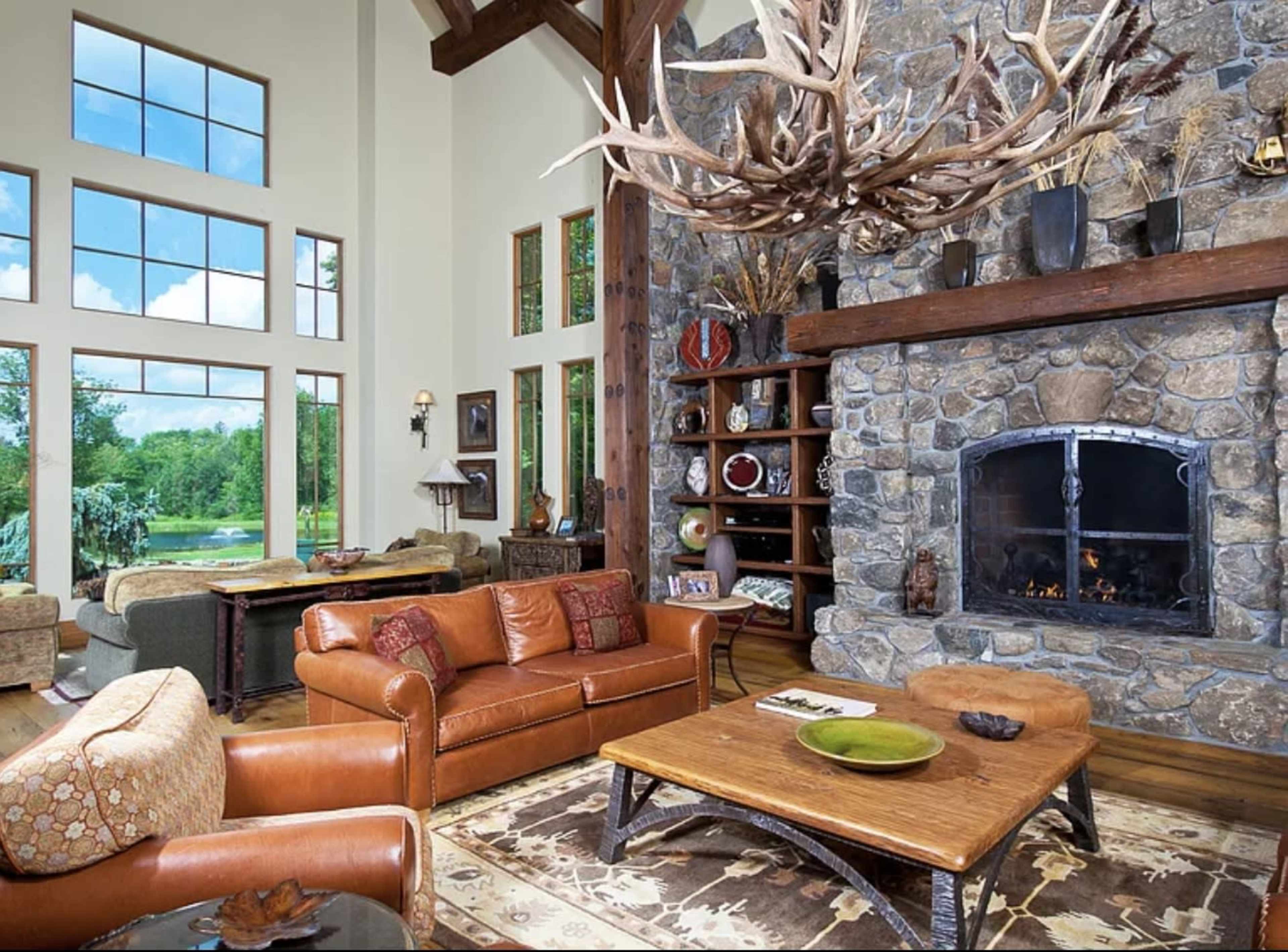 The spacious living room features a stone fireplace, leather seating, and large windows overlooking a green landscape.