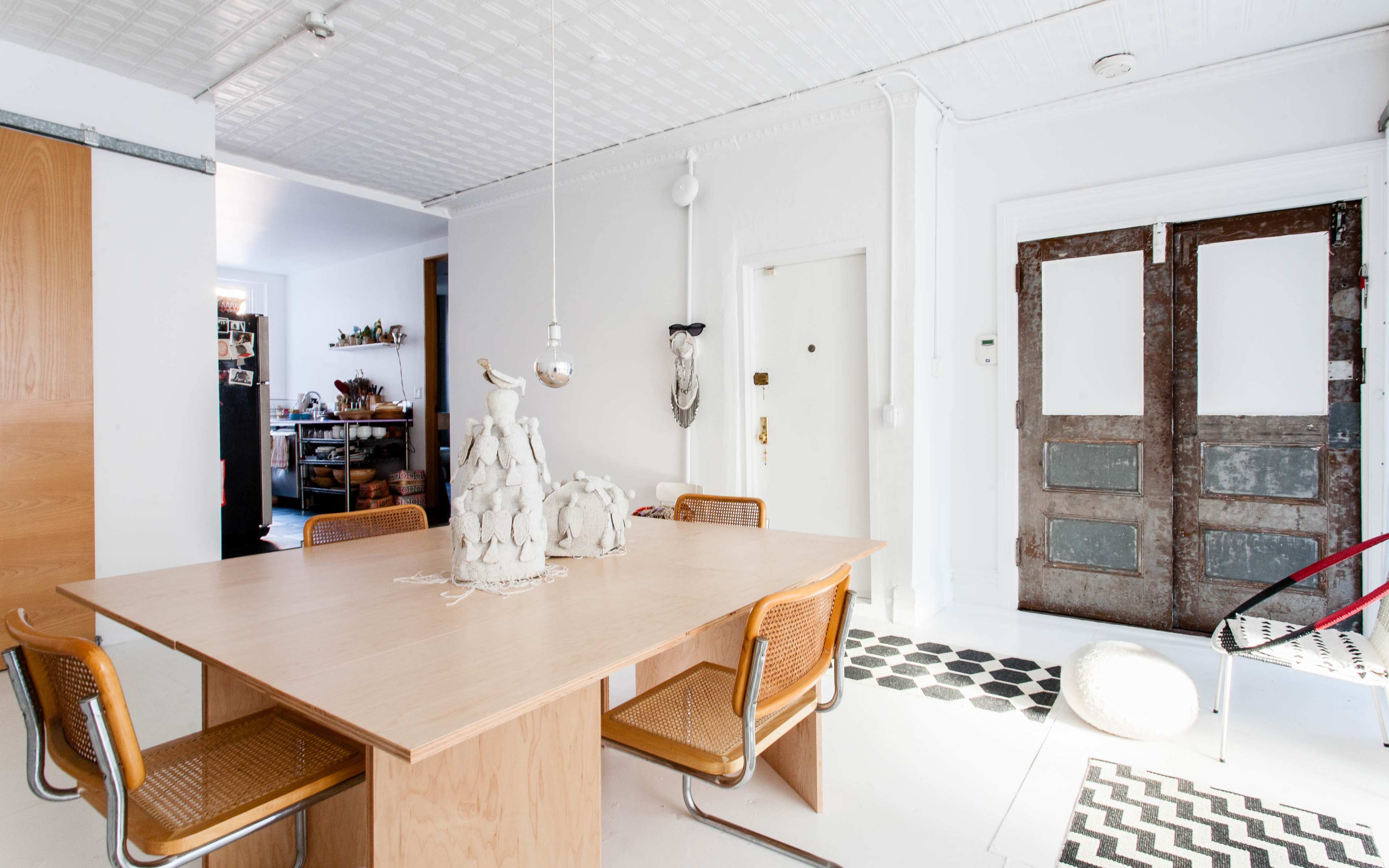 A wooden dining table with four cane chairs is positioned in a bright room featuring a large door with a rustic finish and a patterned rug on the floor.