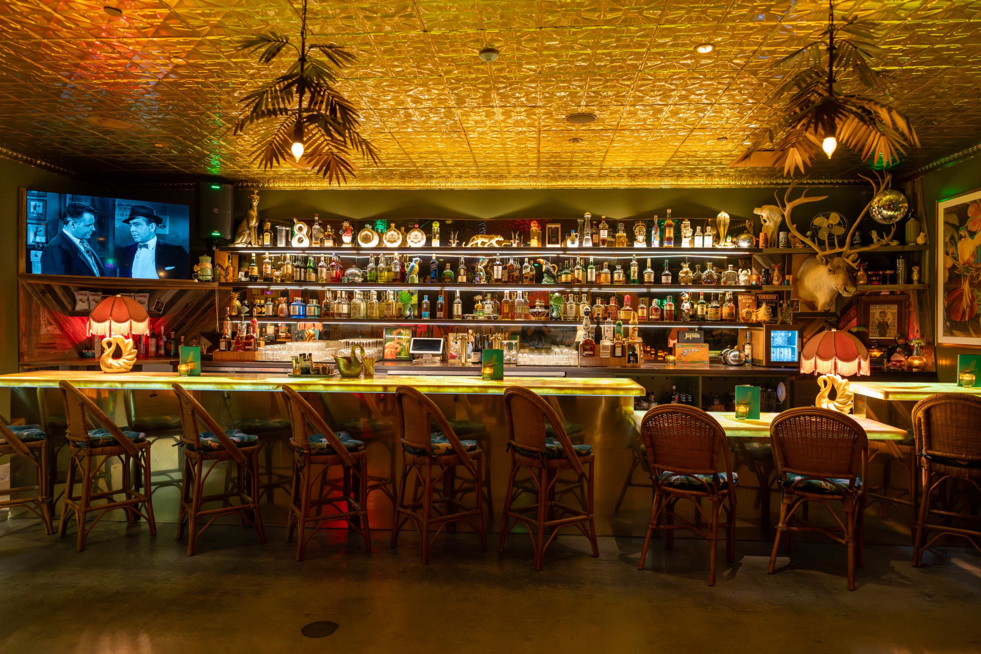 The image shows a vibrant bar interior with a long counter, numerous bottles displayed on shelves, and ambient lighting, including ceiling decorations.