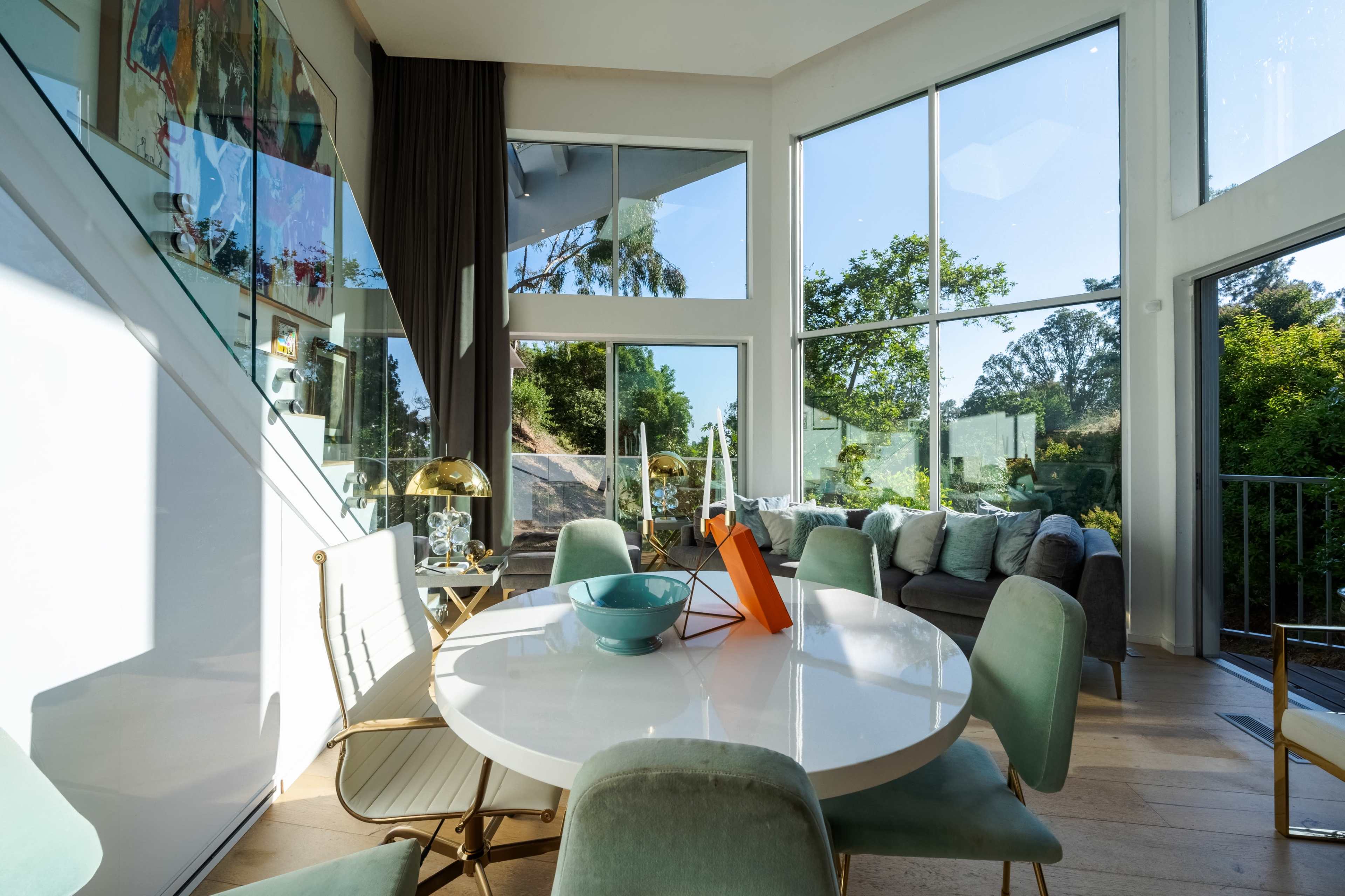 A bright living area features large windows, a round white dining table, and a green sofa, with greenery visible outside.